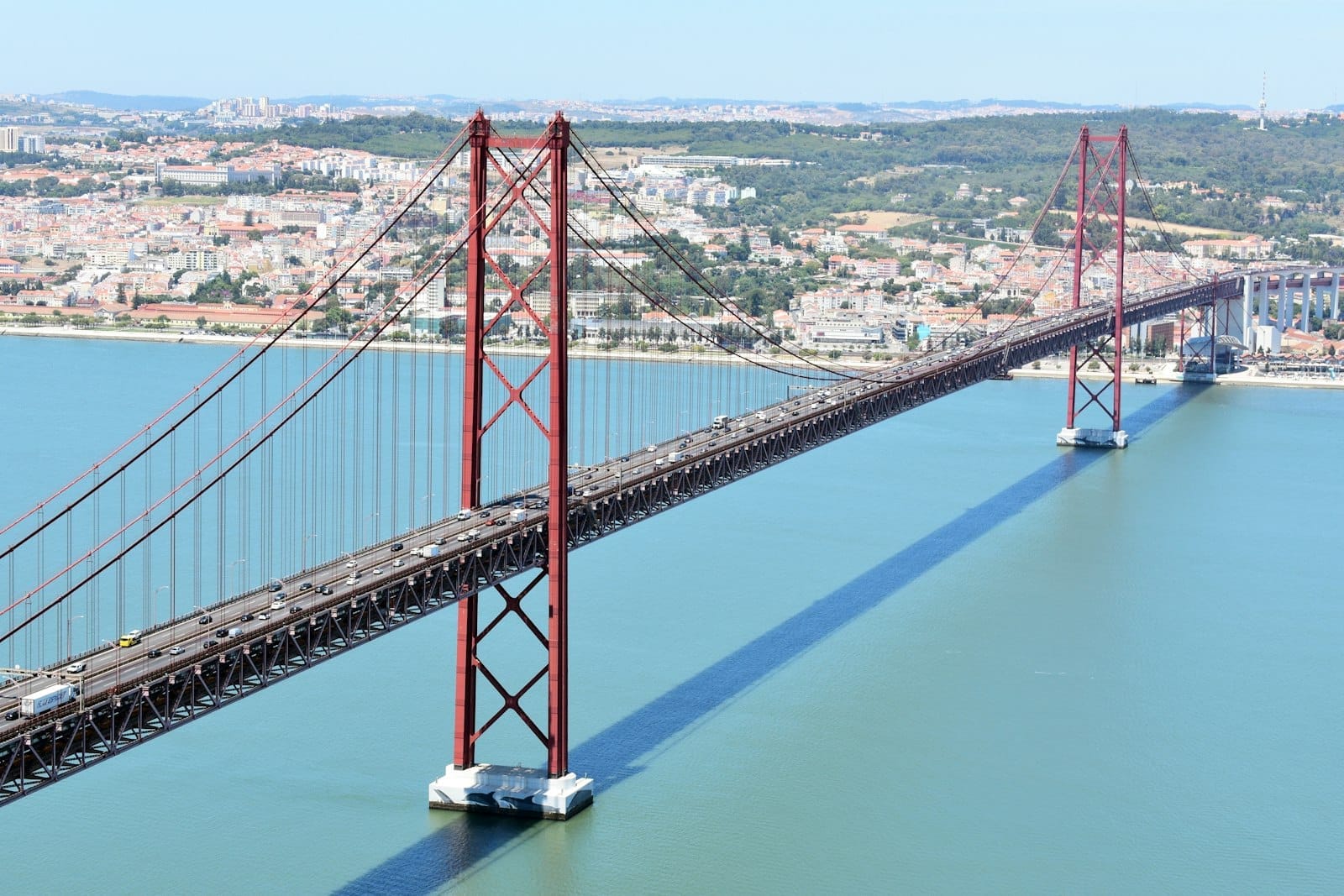Vista aérea da icônica Ponte 25 de Abril, em vermelho, cruzando o Rio Tejo em Lisboa, com a cidade e colinas verdes ao fundo.