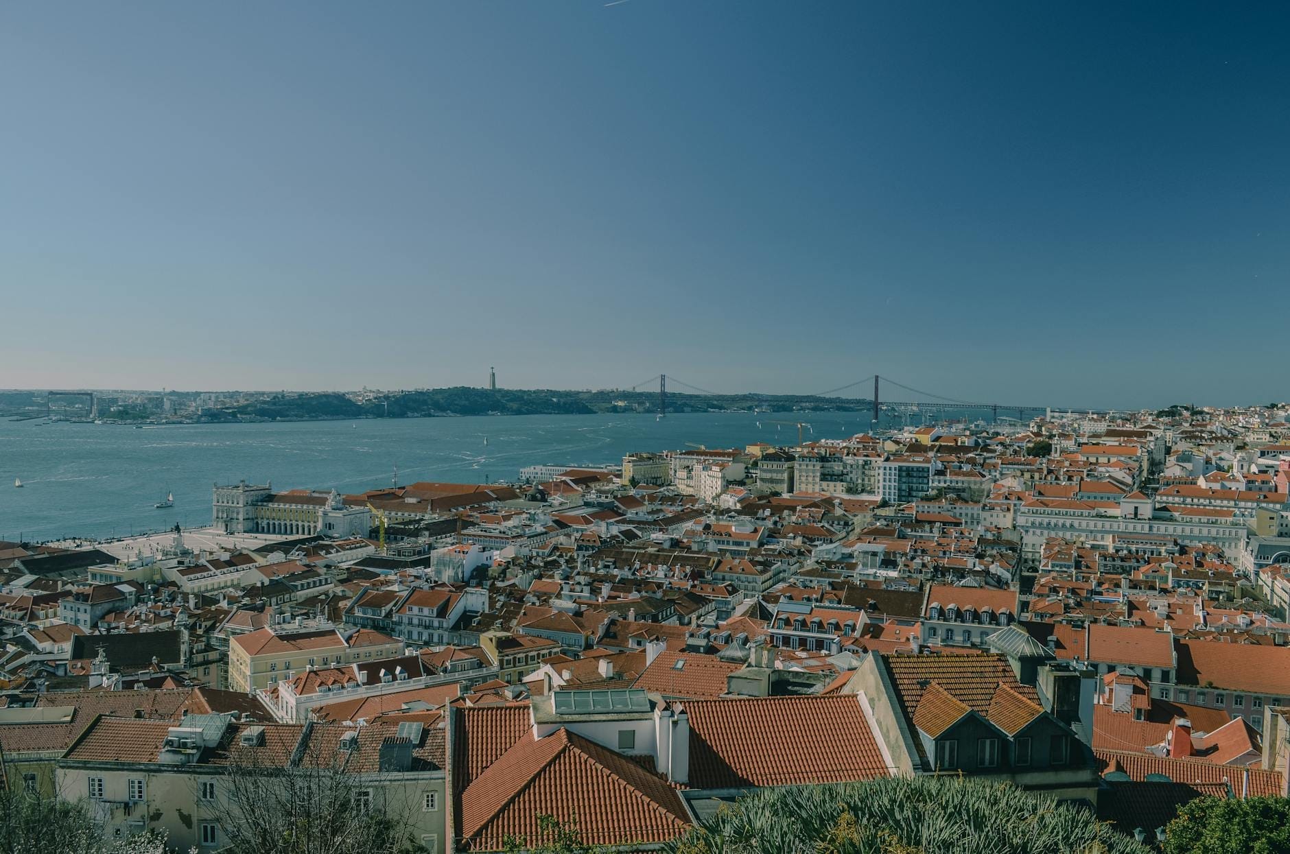 Large vue sur les toits rouges de Lisbonne avec le Tage et le Pont du 25 de Abril visibles en arrière-plan sous un ciel bleu dégagé.