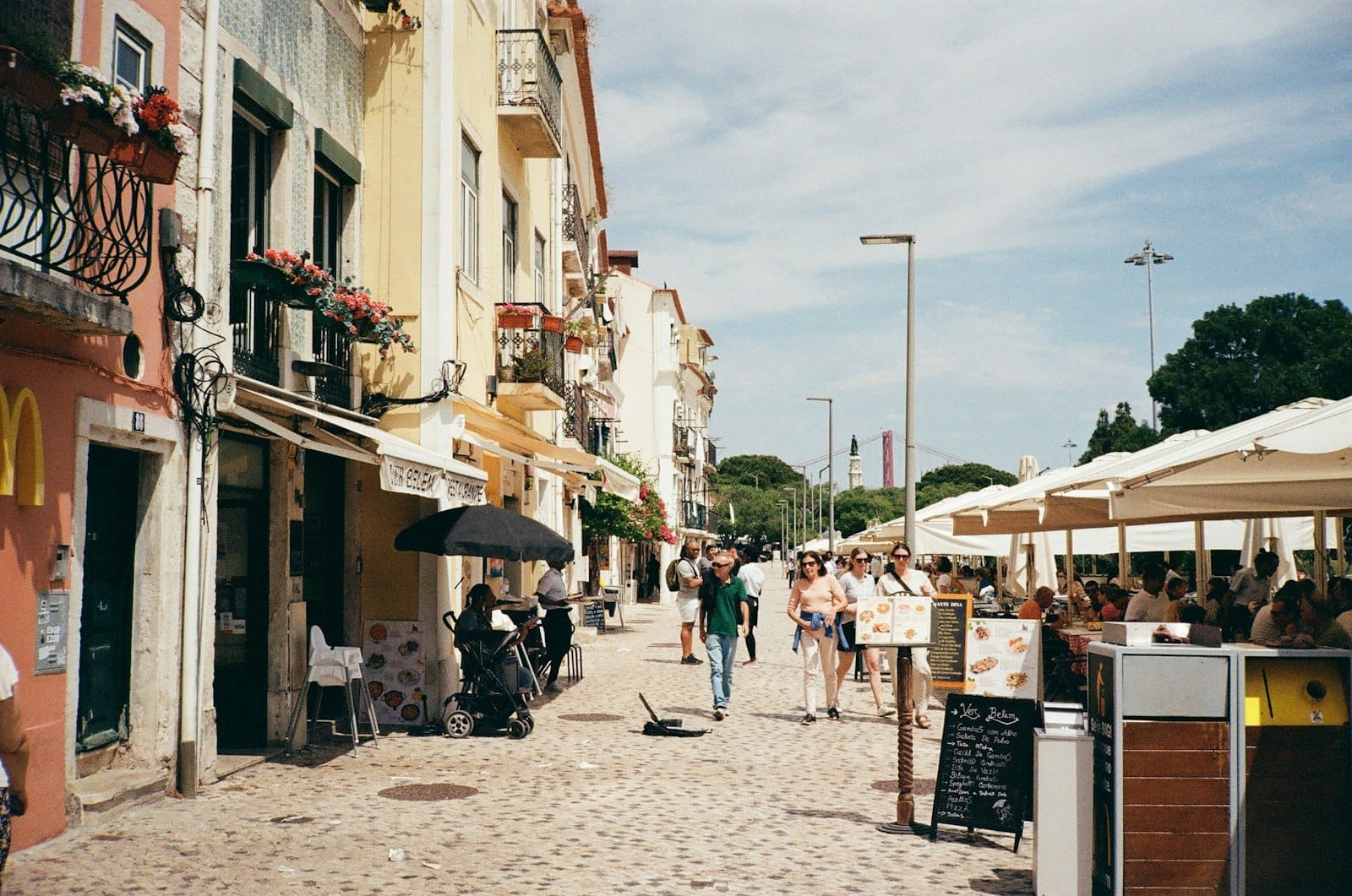 Restaurantes ao ar livre com pessoas jantando sob guarda-sóis em uma animada rua de paralelepípedos em Lisboa num dia de sol.