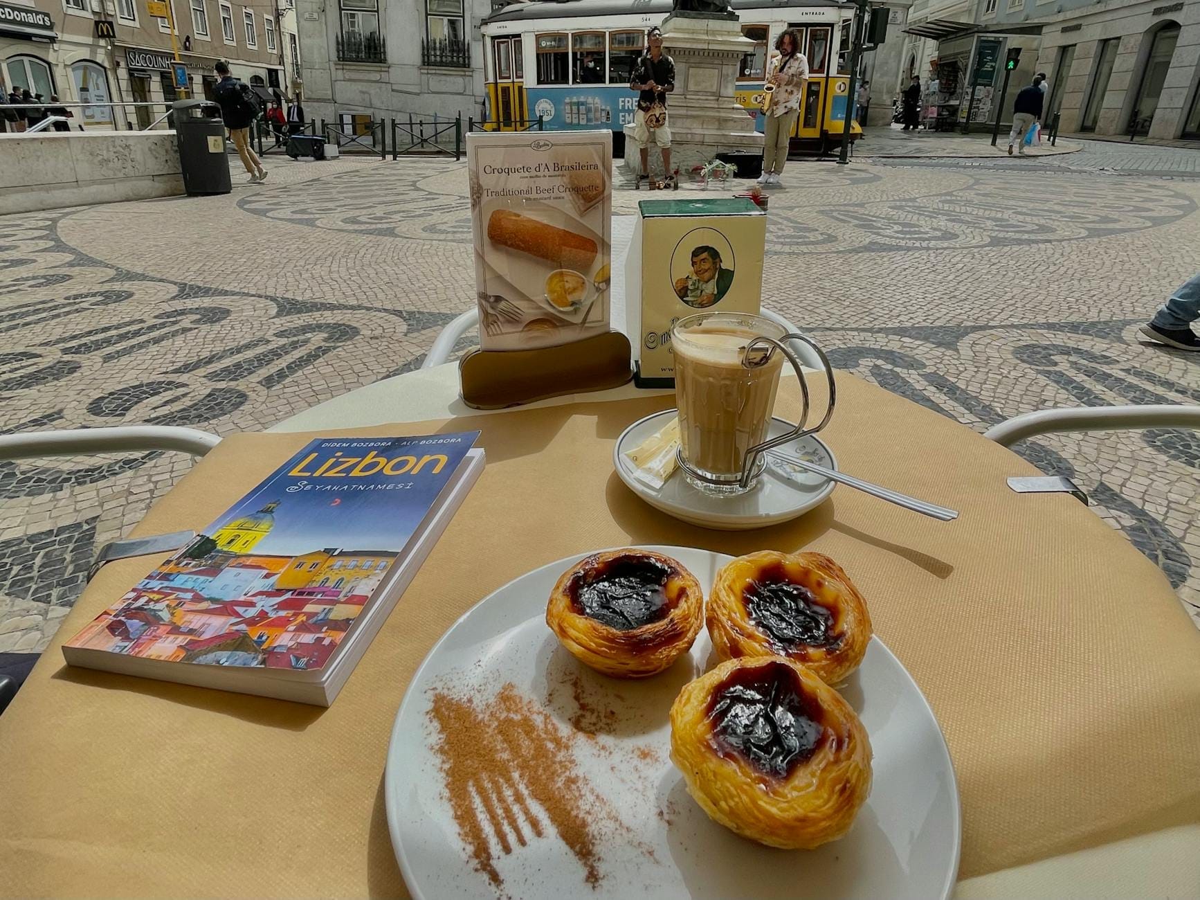 Mesa com três pasteis de nata, café e um guia de Lisboa numa esplanada com elétrico e praça ao fundo.