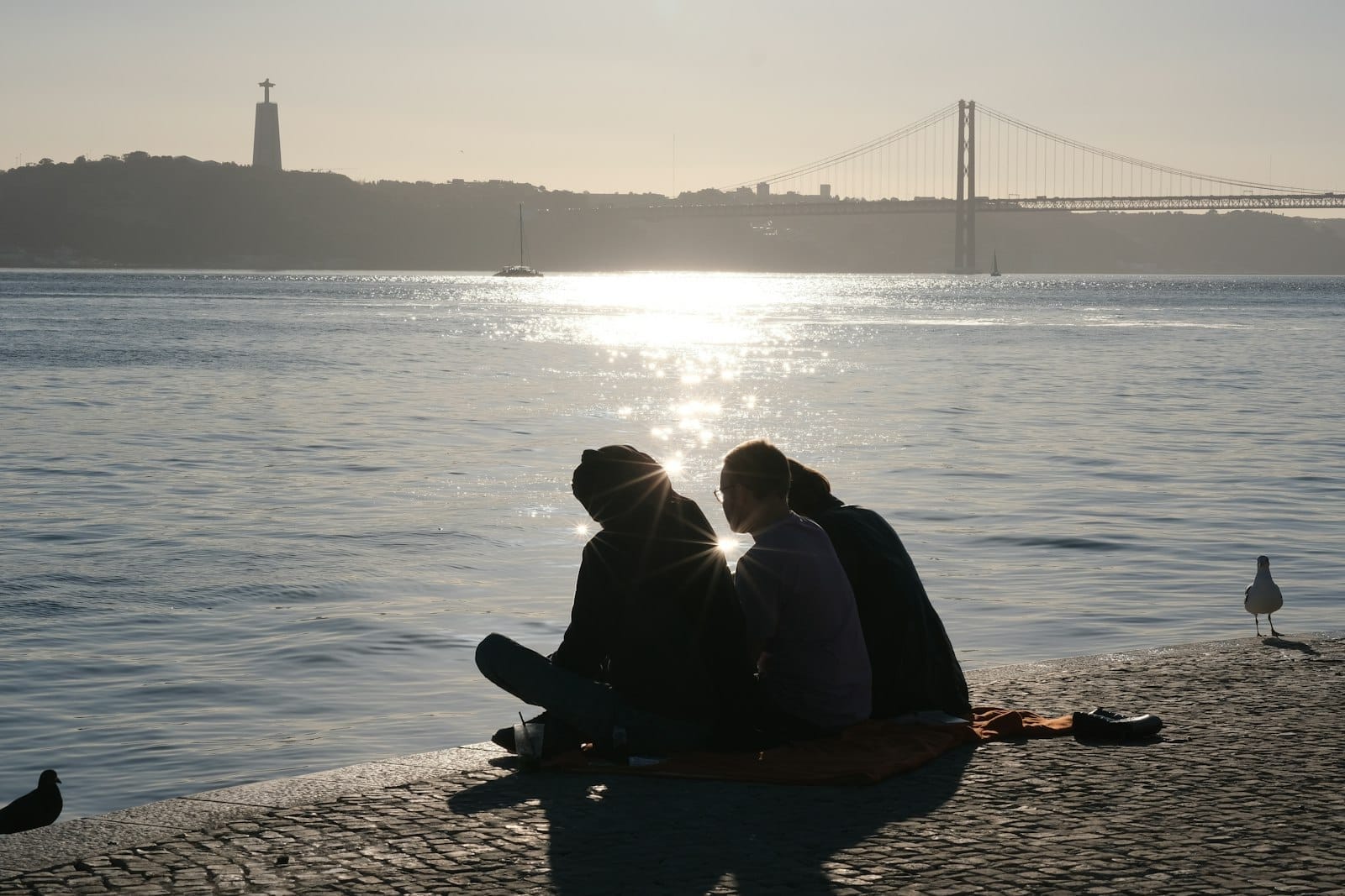 Casal sentado juntinho à beira d'água ao pôr do sol em Lisboa, com a Ponte 25 de Abril e o Cristo Rei ao fundo.