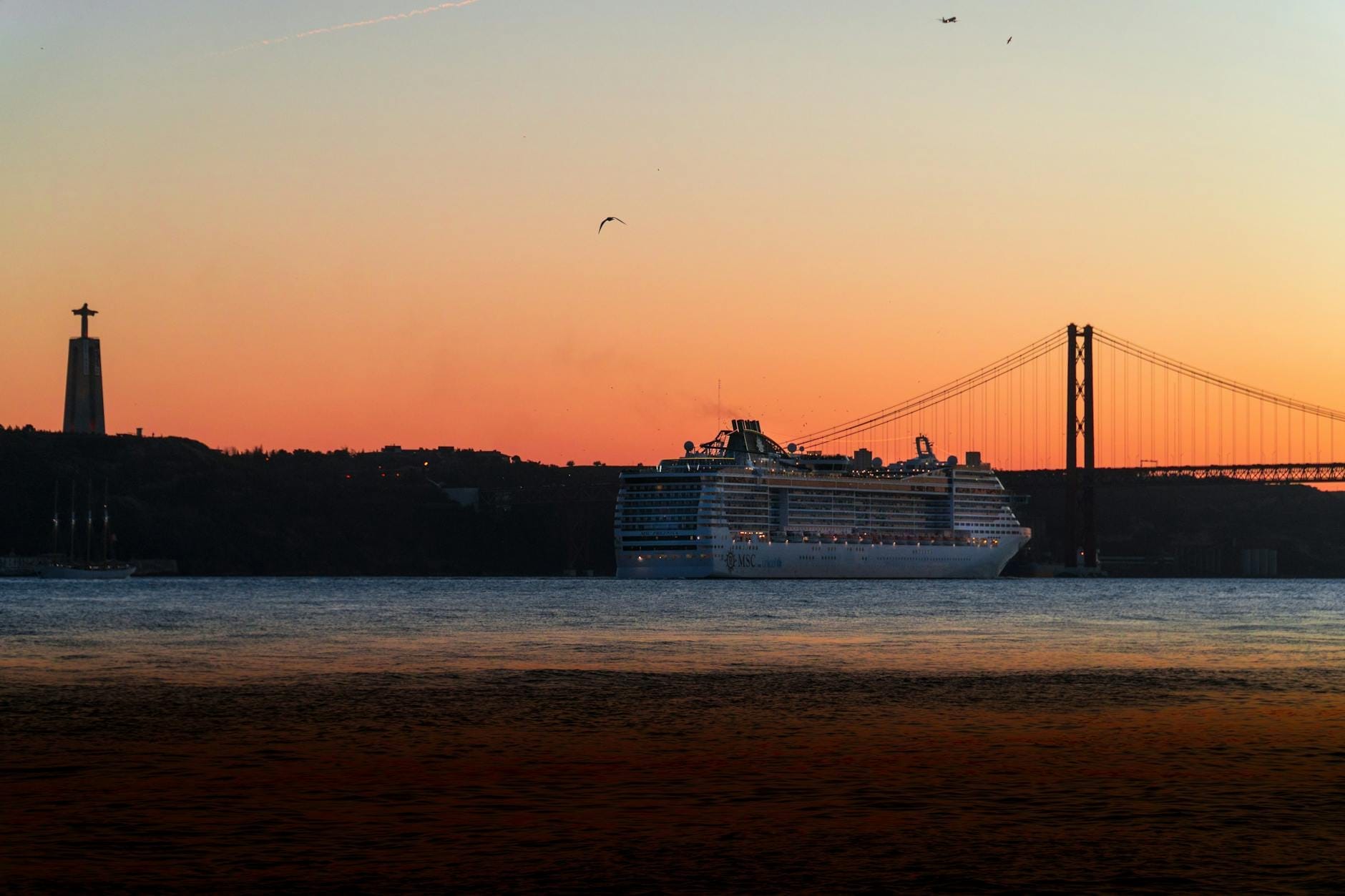Un grand bateau de croisière sur le Tage au coucher du soleil avec le Pont 25 de Abril et la statue du Cristo Rei en arrière-plan.