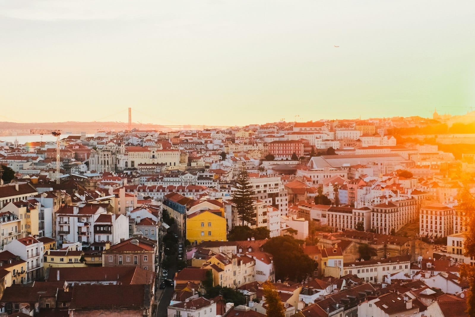Panorama de Lisbonne au coucher du soleil, avec des toits en terre cuite, des bâtiments colorés et une douce lumière dorée illuminant la ligne d'horizon et les monuments lointains.
