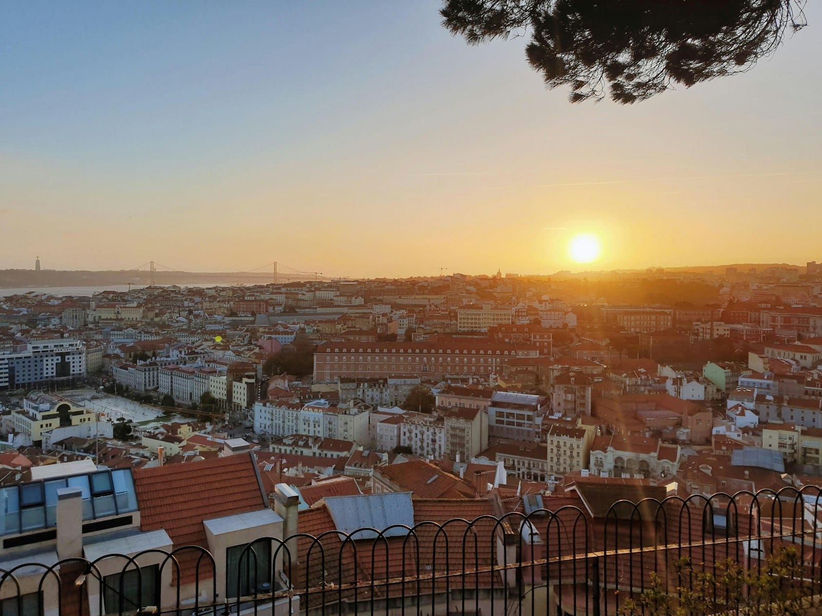 Vue du coucher de soleil sur les toits de Lisbonne et le Tage, vue depuis un belvédère élevé avec une clôture en fer et des branches d'arbres au-dessus.