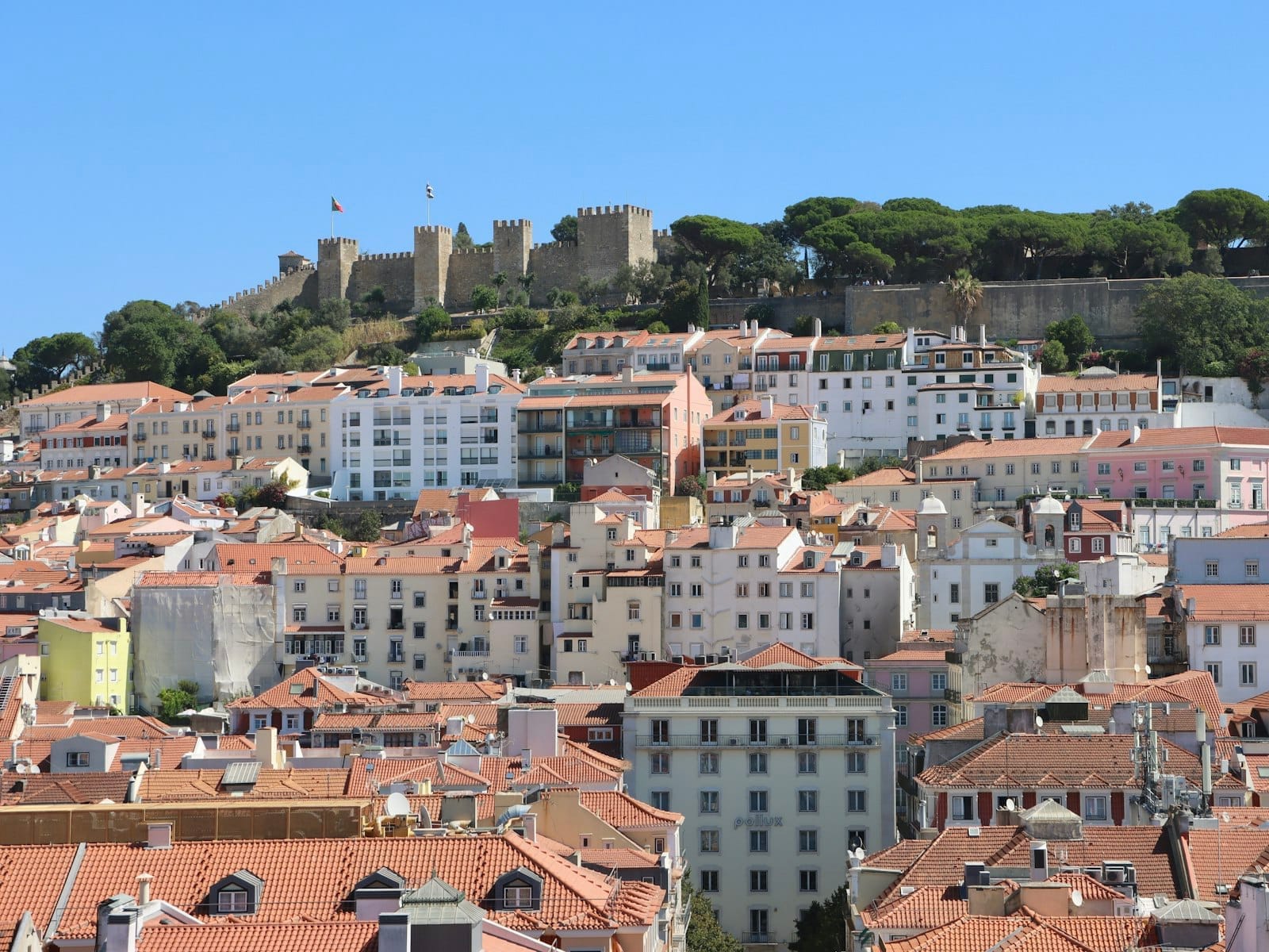 Vue panoramique du quartier Alfama de Lisbonne avec le Castelo de São Jorge au sommet de la colline, bâtiments historiques et verdure luxuriante sous un ciel bleu dégagé.