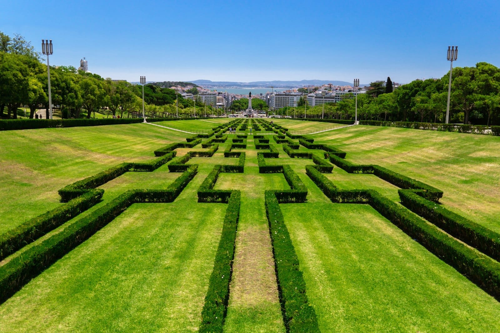 Jardins symétriques luxuriants et haies taillées dans le Parc Eduardo VII de Lisbonne par une journée ensoleillée, avec des vues sur la ville en arrière-plan.