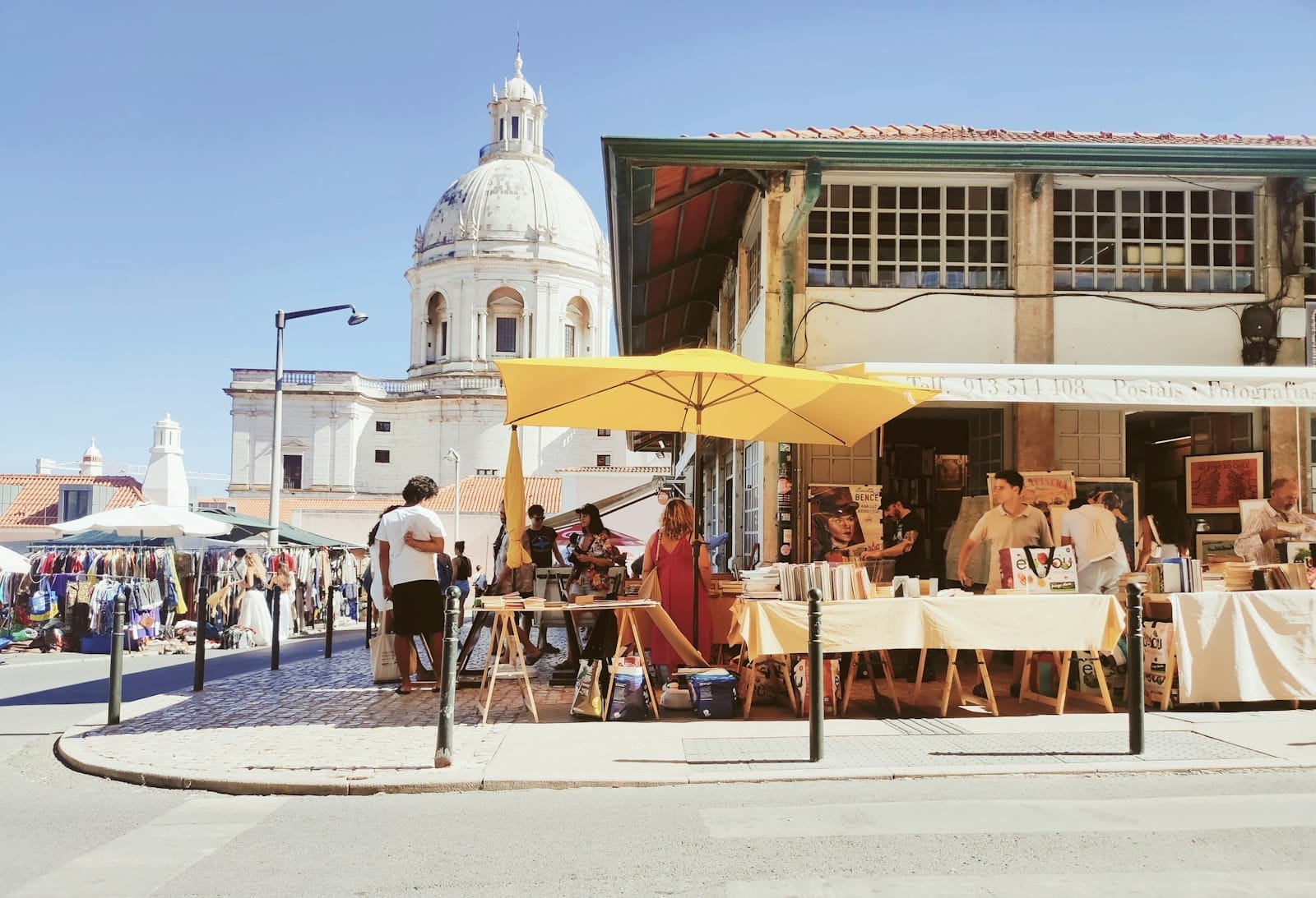 Scène de marché de rue en plein air à Lisbonne avec des étals sous des parasols jaunes, des gens qui flânent et une église à dôme en arrière-plan.