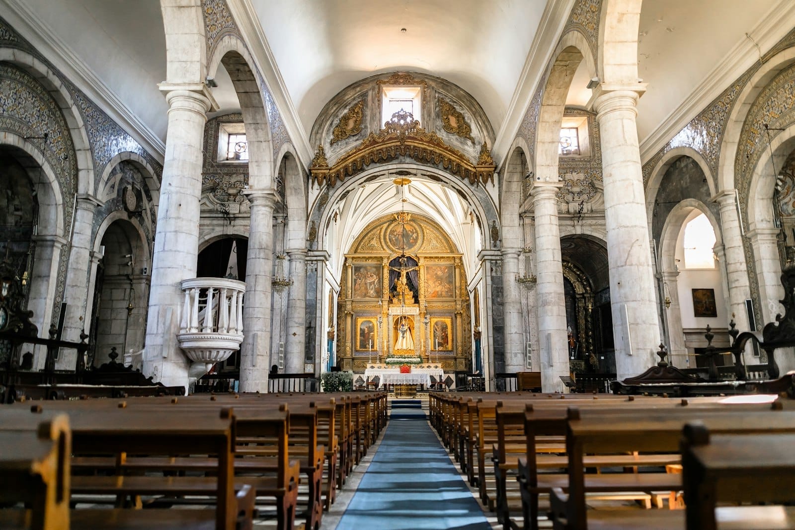 Intérieur bien éclairé d'une église historique en pierre à Lisbonne avec des plafonds voûtés, des colonnes classiques, des bancs en bois et un riche retable doré à l'avant.