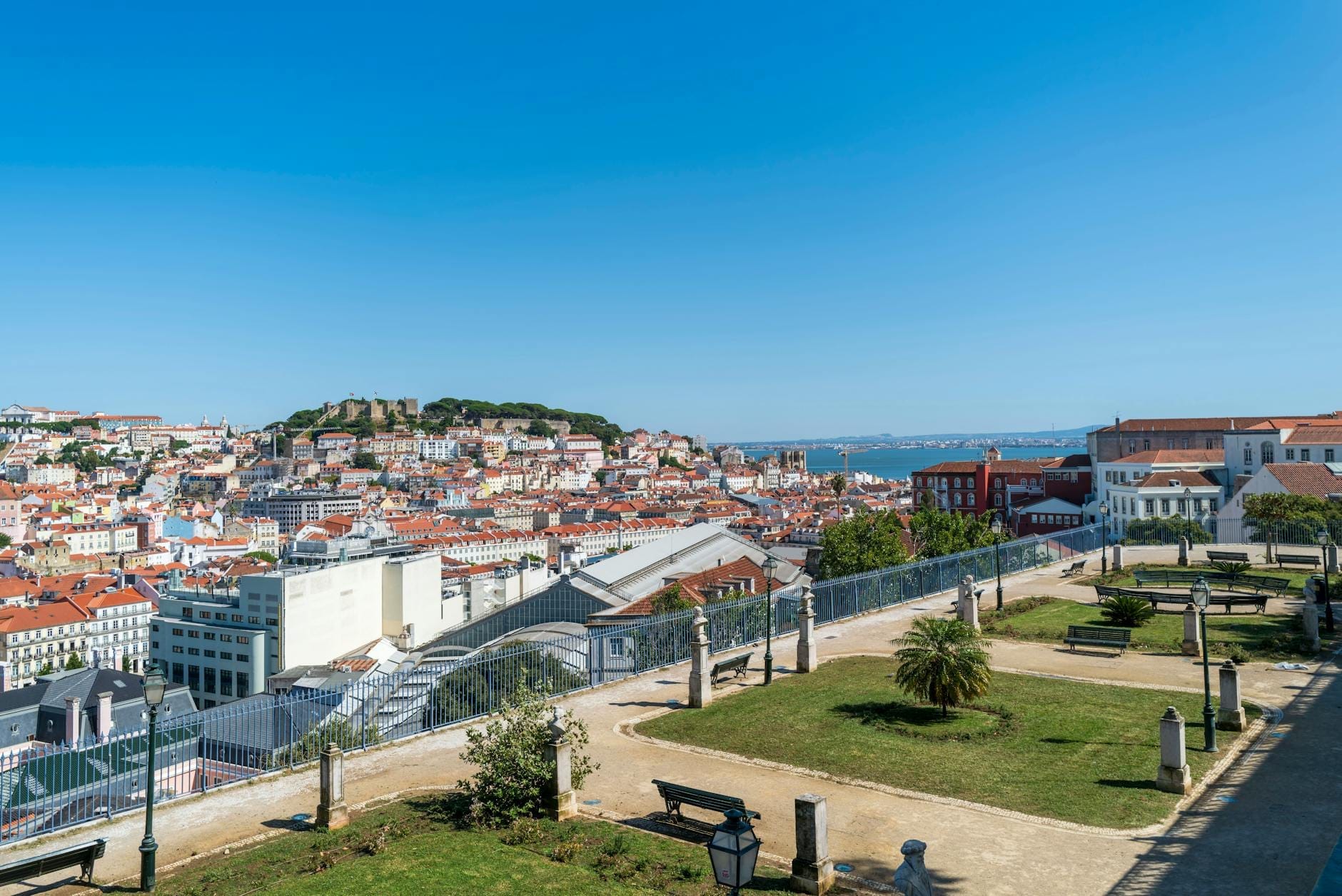 Paysage urbain de Lisbonne sous un ciel bleu dégagé avec un soleil éclatant, montrant des toits rouges, des collines lointaines et le Tage par une sèche journée d'été.