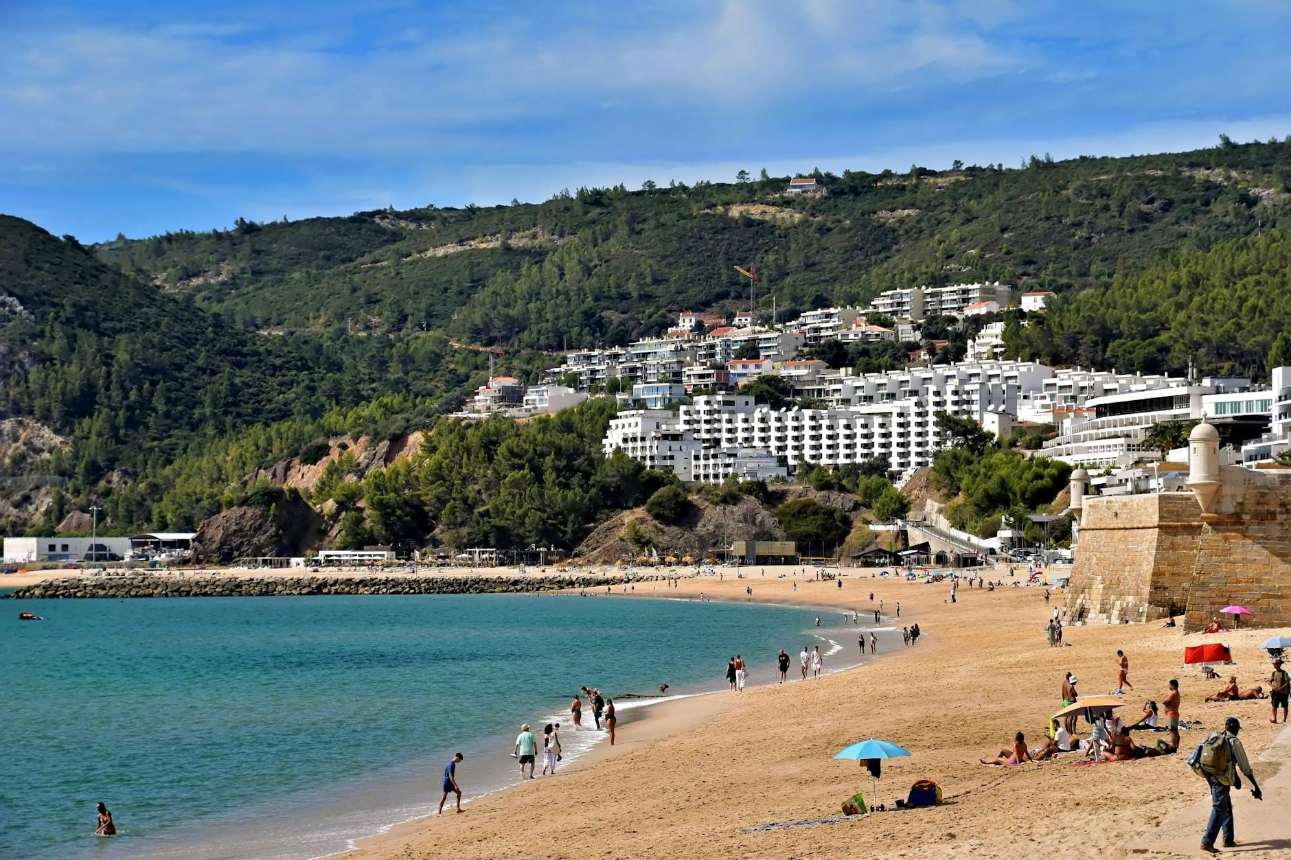 Des personnes profitant d'une plage de sable aux eaux turquoise calmes, avec des parasols et un arrière-plan de bâtiments blancs et de collines verdoyantes près de Lisbonne.
