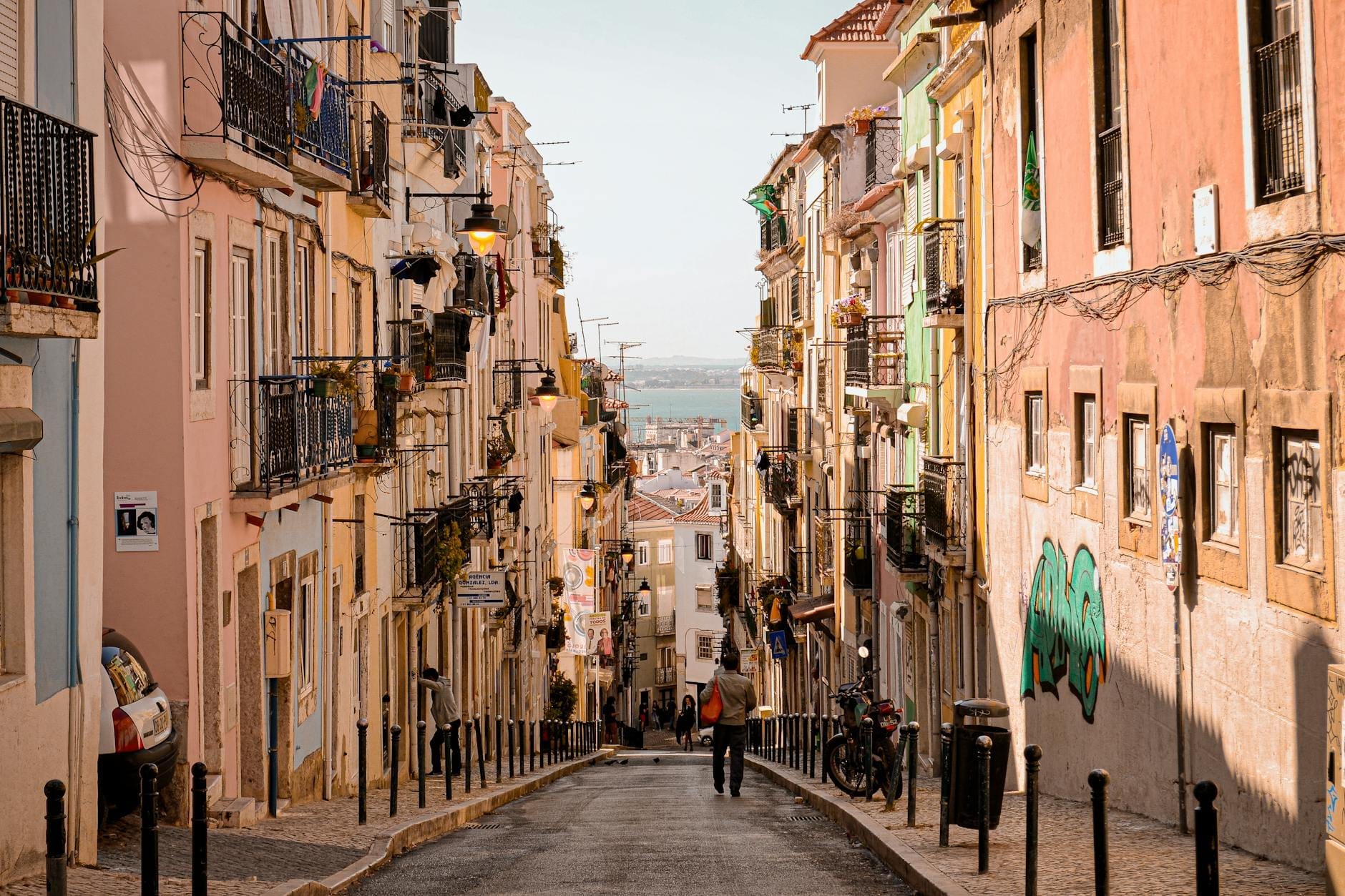 Rue étroite et escarpée bordée de bâtiments traditionnels colorés de Lisbonne, personnes marchant en descente et vue lointaine sur le paysage urbain et l'eau.