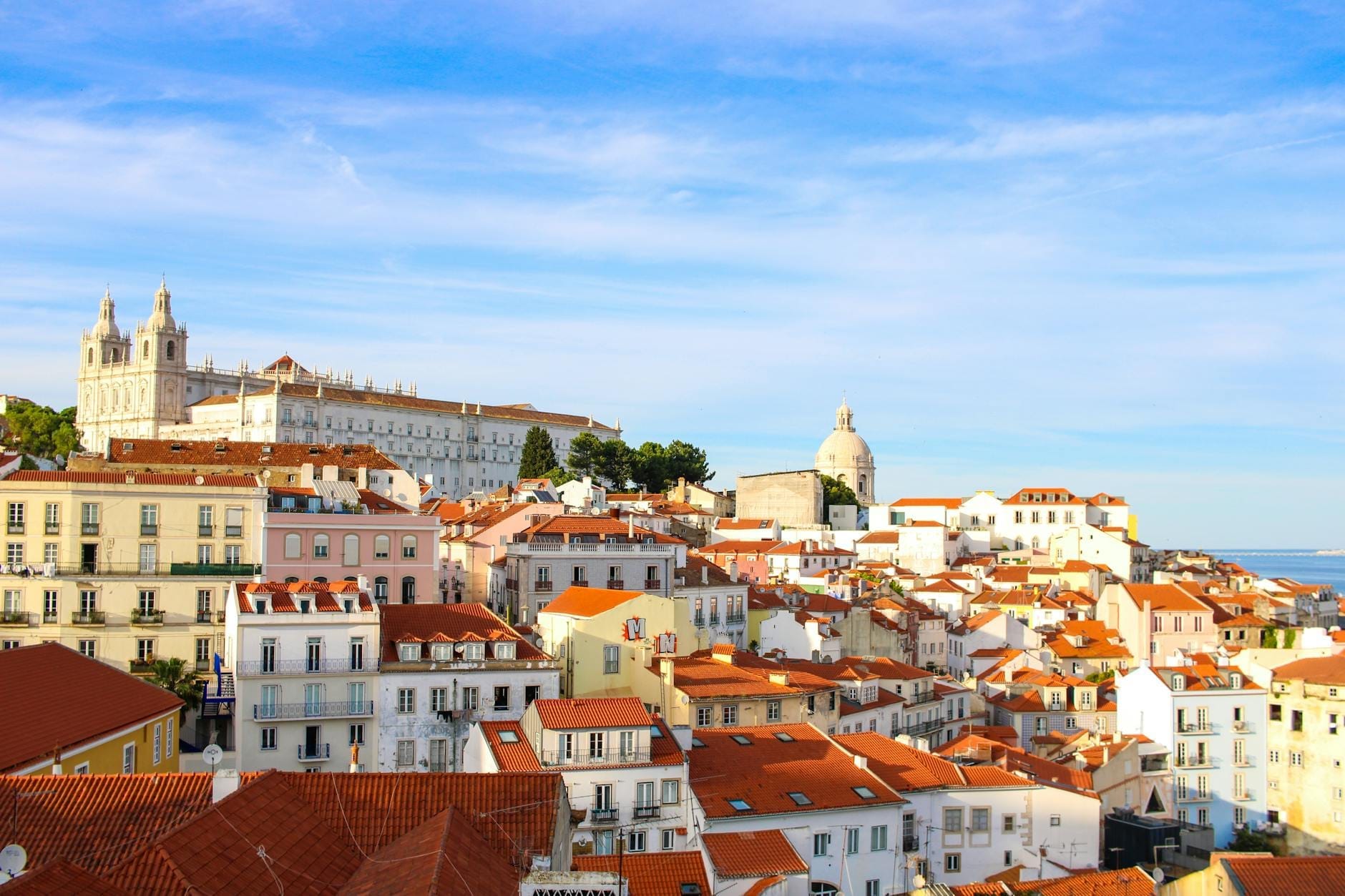 Vista panorâmica luminosa e ensolarada do histórico bairro de Alfama em Lisboa, com telhados vermelhos, grandes edifícios brancos e um céu azul, captando o charme e a elegância da cidade.