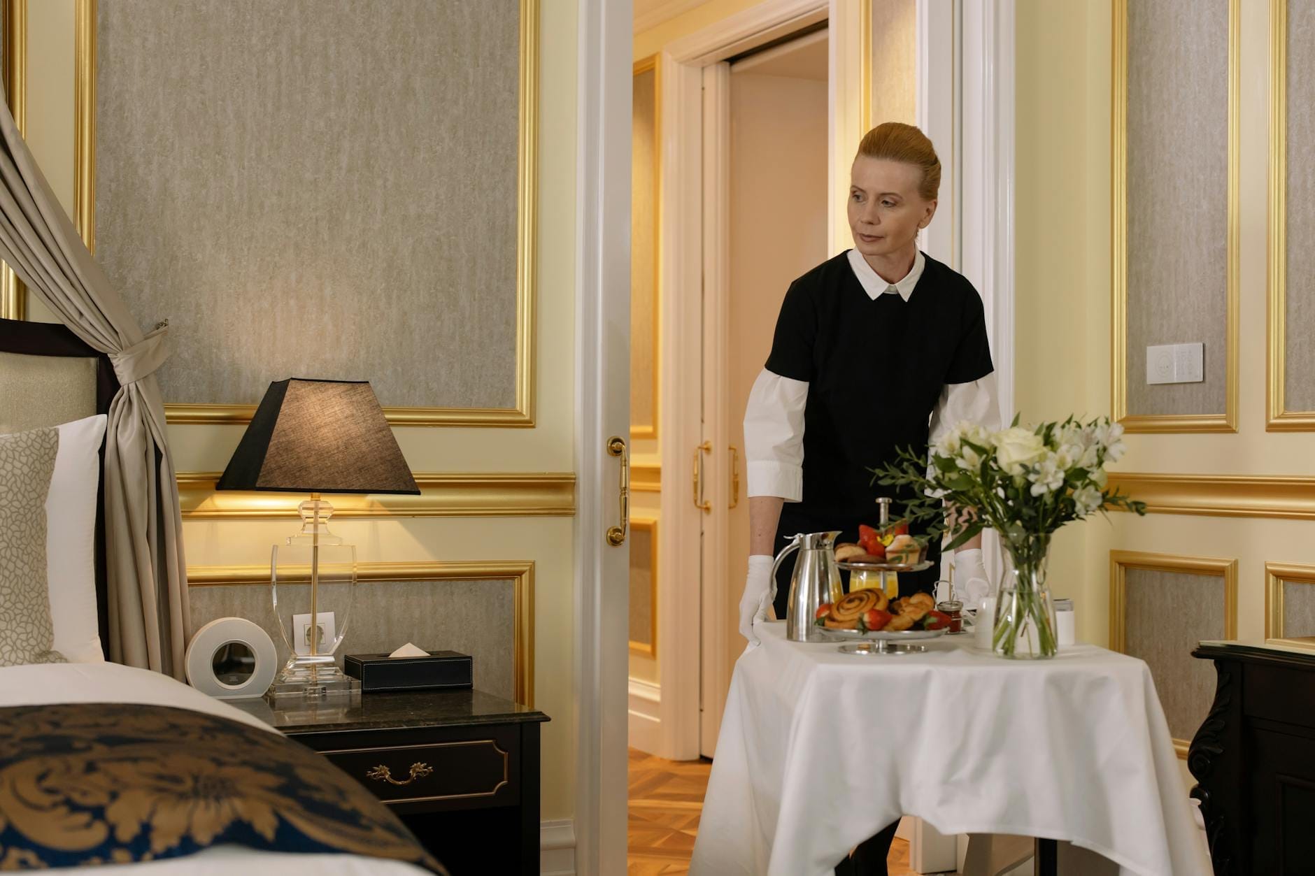 Uma suíte de hotel luxuosa com elegantes detalhes dourados, um funcionário bem-vestido servindo café da manhã em uma mesinha dobrável e uma atmosfera aconchegante e sofisticada.