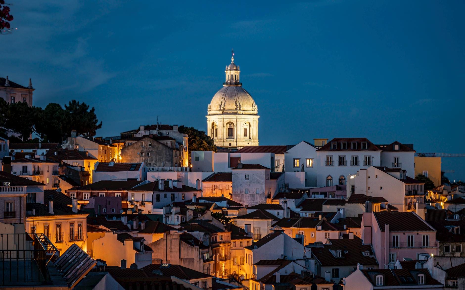 Le quartier d'Alfama à Lisbonne la nuit avec un dôme illuminé, des bâtiments blanchis à la chaux et des toits orangés sous un ciel bleu profond.