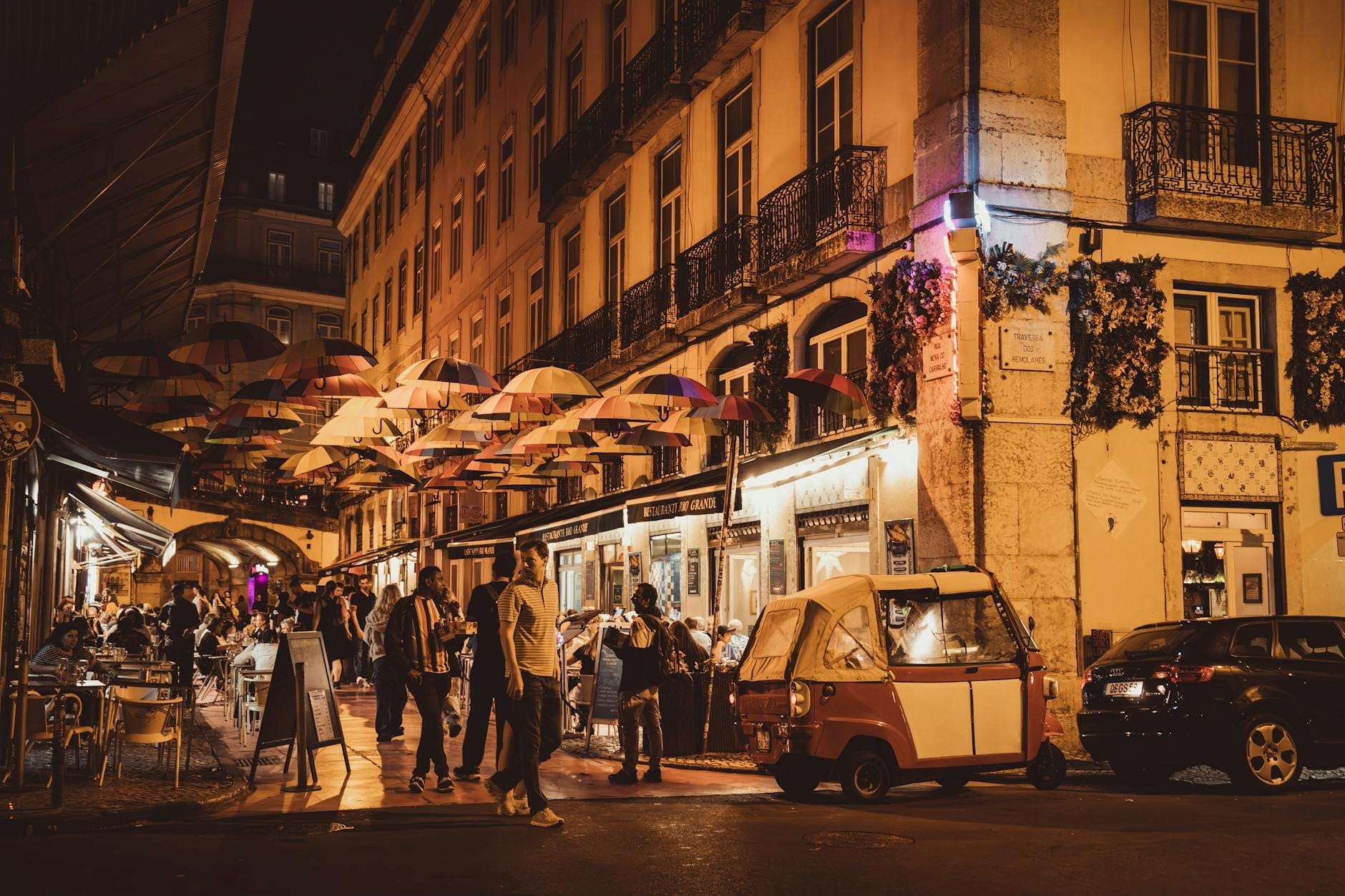 Une rue animée de Lisbonne la nuit avec des gens devant les bars, des bâtiments historiques illuminés et des parapluies colorés suspendus au-dessus.