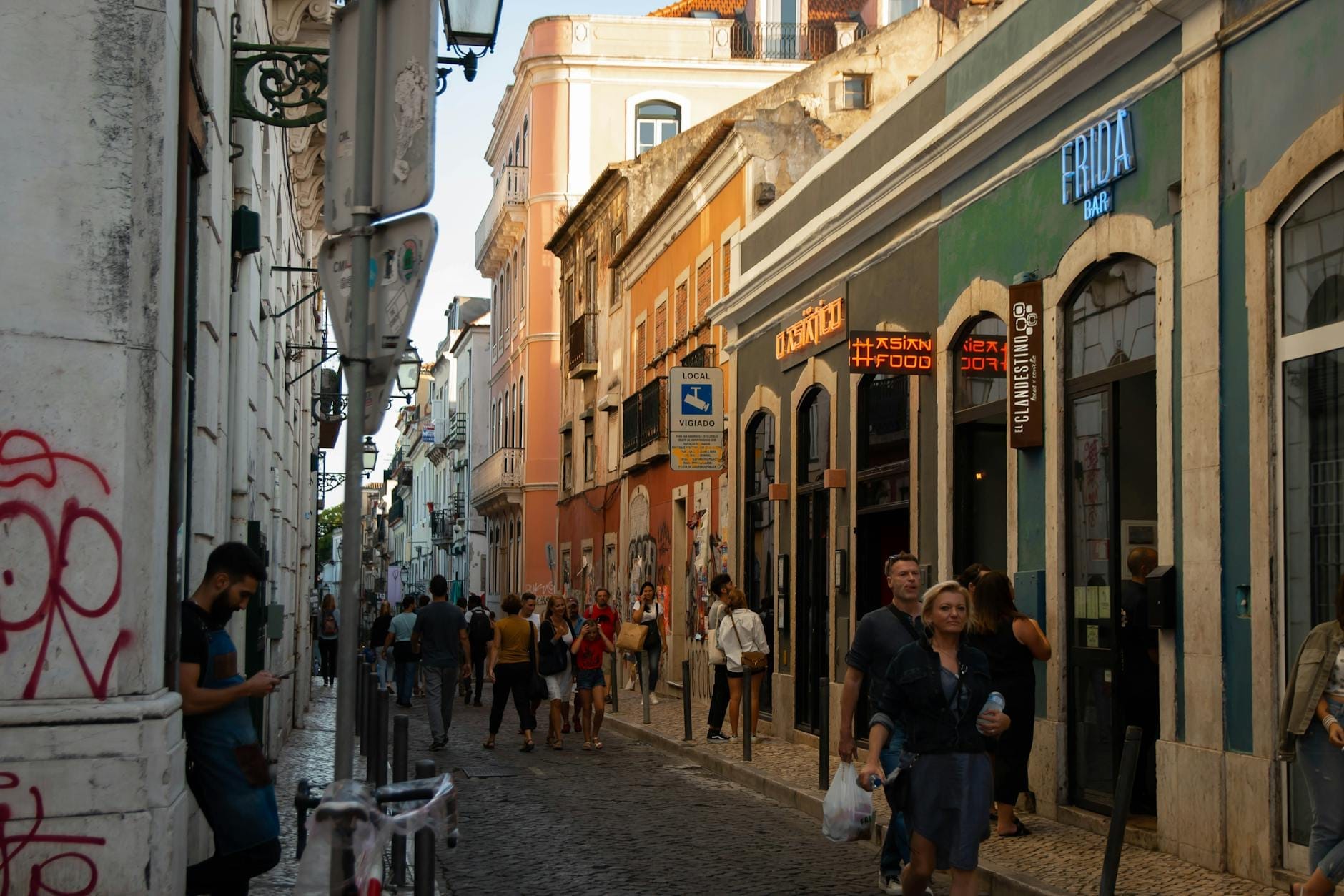 Rue étroite à Bairro Alto, Lisbonne, bordée de bars et de personnes se promenant et socialisant en soirée.