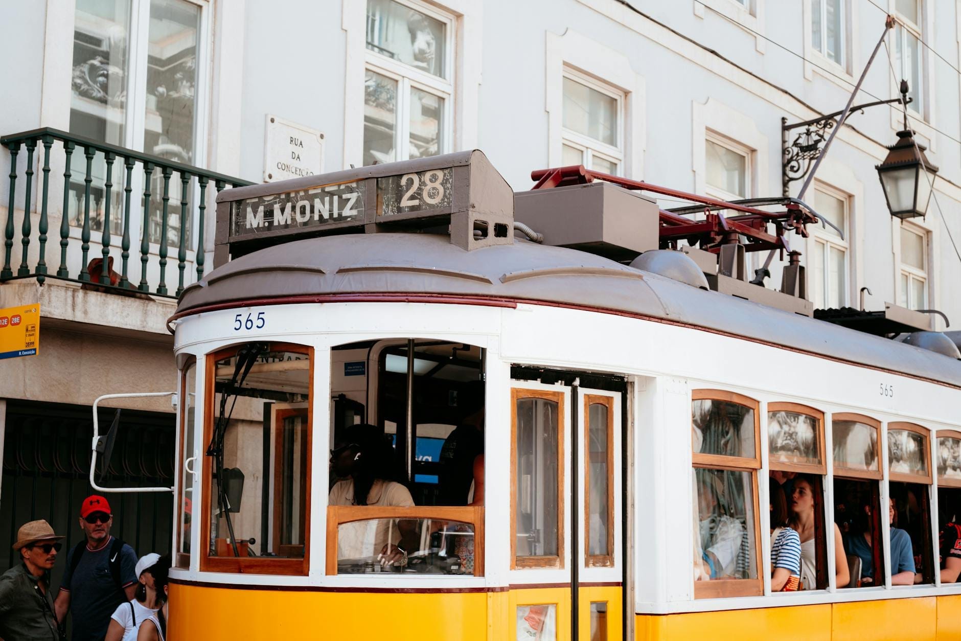Tramway jaune classique numéro 28 rempli de passagers circulant dans une rue typique de Lisbonne avec des bâtiments blancs et des personnes attendant à proximité.