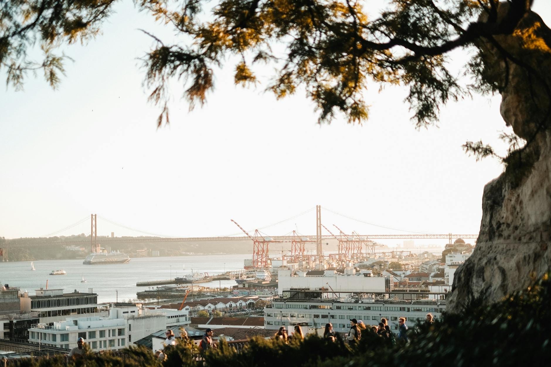 Multidões reunidas num miradouro de Lisboa com vista para o rio Tejo, com a Ponte 25 de Abril e a luz do verão ao fundo.