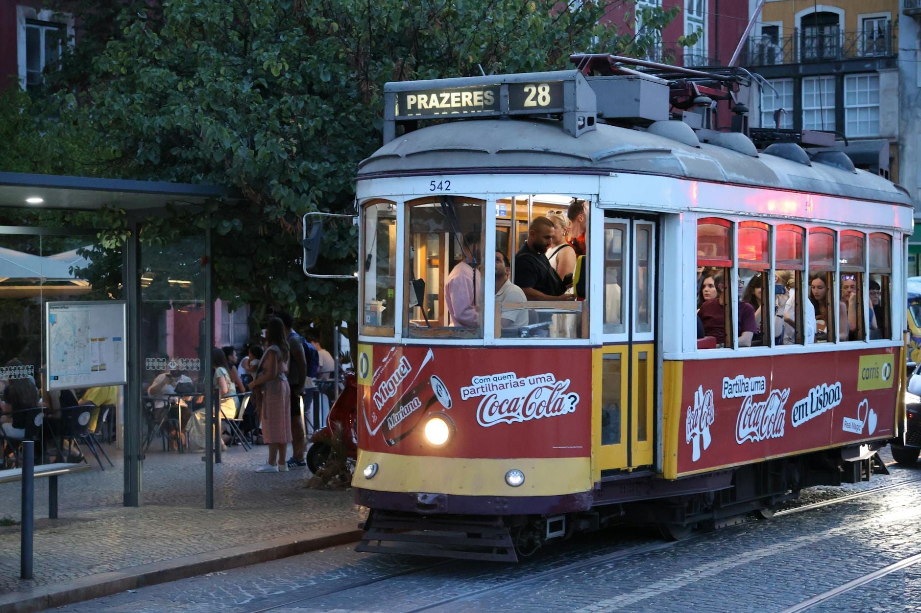 Elétrico 28 de Lisboa lotado parado numa estação urbana, cheio de passageiros, com pessoas à espera nas proximidades e edifícios clássicos ao fundo.