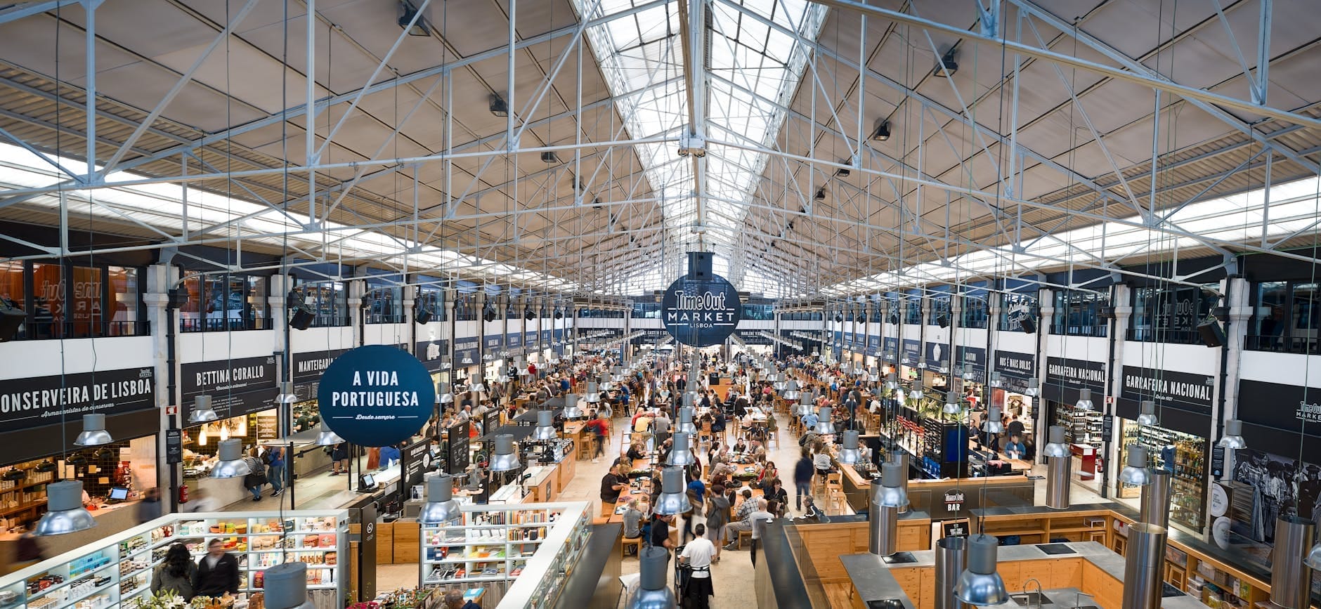 Mercado coberto movimentado em Lisboa, com pessoas comprando em várias bancas sob um teto de vidro alto, mostrando a atmosfera comercial vibrante da cidade.