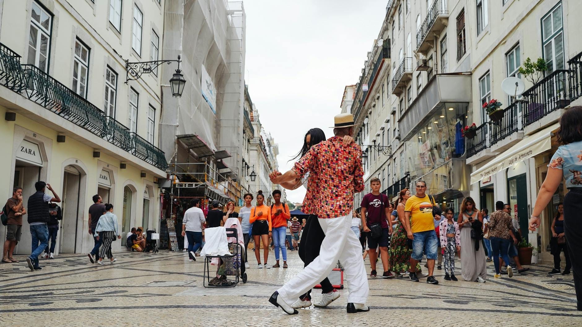 Large rue piétonne commerçante à Lisbonne bordée de boutiques, de personnes se promenant et faisant leurs achats, et un couple dansant au premier plan.