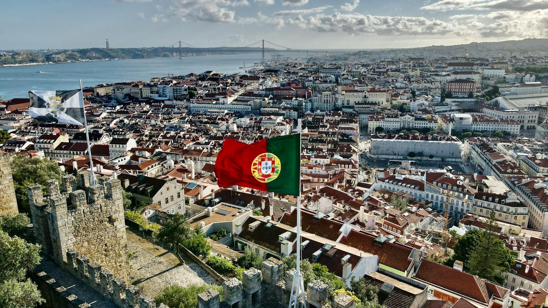 Large vue aérienne de Lisbonne avec le drapeau portugais et les emblématiques toits rouges, le fleuve et les monuments de la ville visibles en arrière-plan.