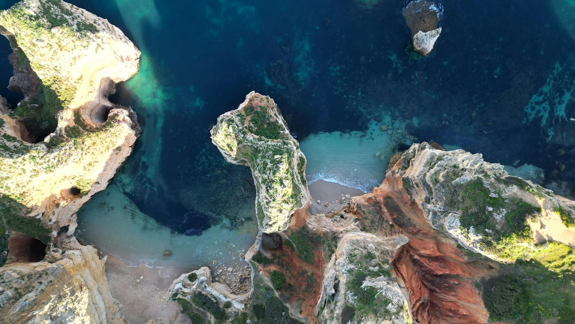 Vista aérea do dramático litoral do Algarve perto de Lagos, com água azul cristalina, falésias imponentes, enseadas de areia e vegetação exuberante sob um sol brilhante.