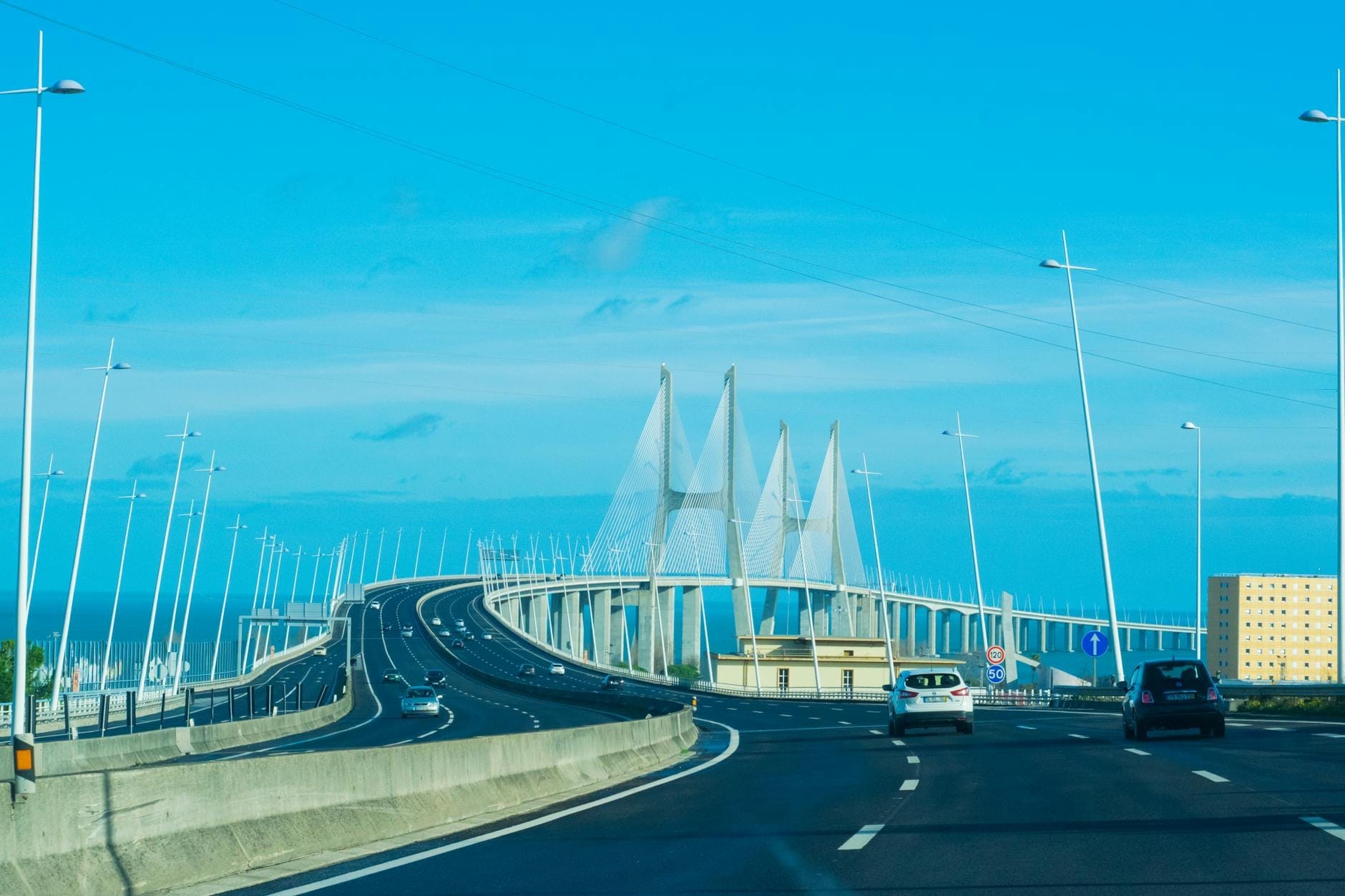 Carros atravessando a moderna Ponte Vasco da Gama em Lisboa sob um céu azul limpo.