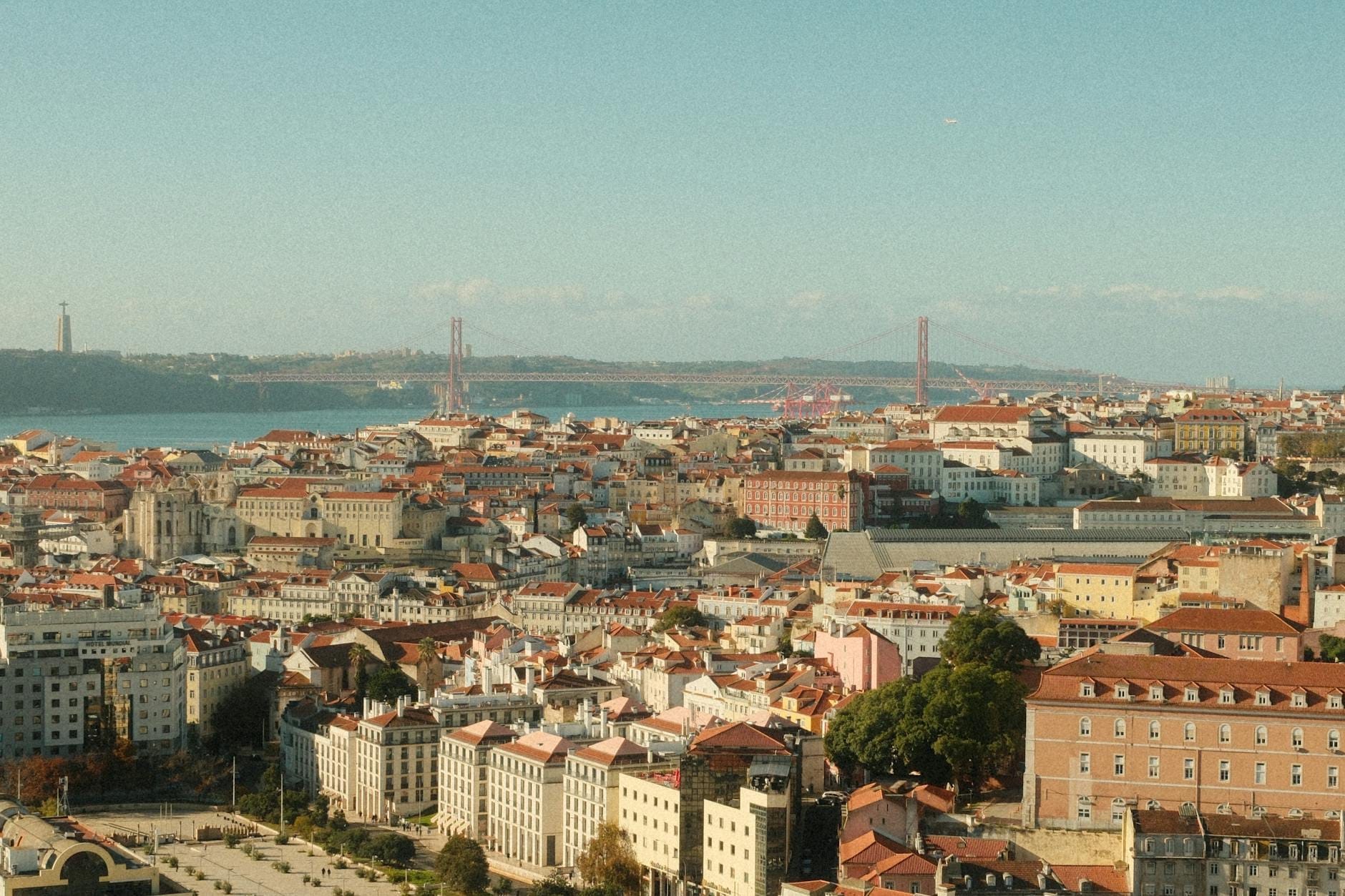 Vista ampla da paisagem urbana de Lisboa com telhados vermelhos, o rio Tejo e a Ponte 25 de Abril sob um céu predominantemente limpo e ensolarado.