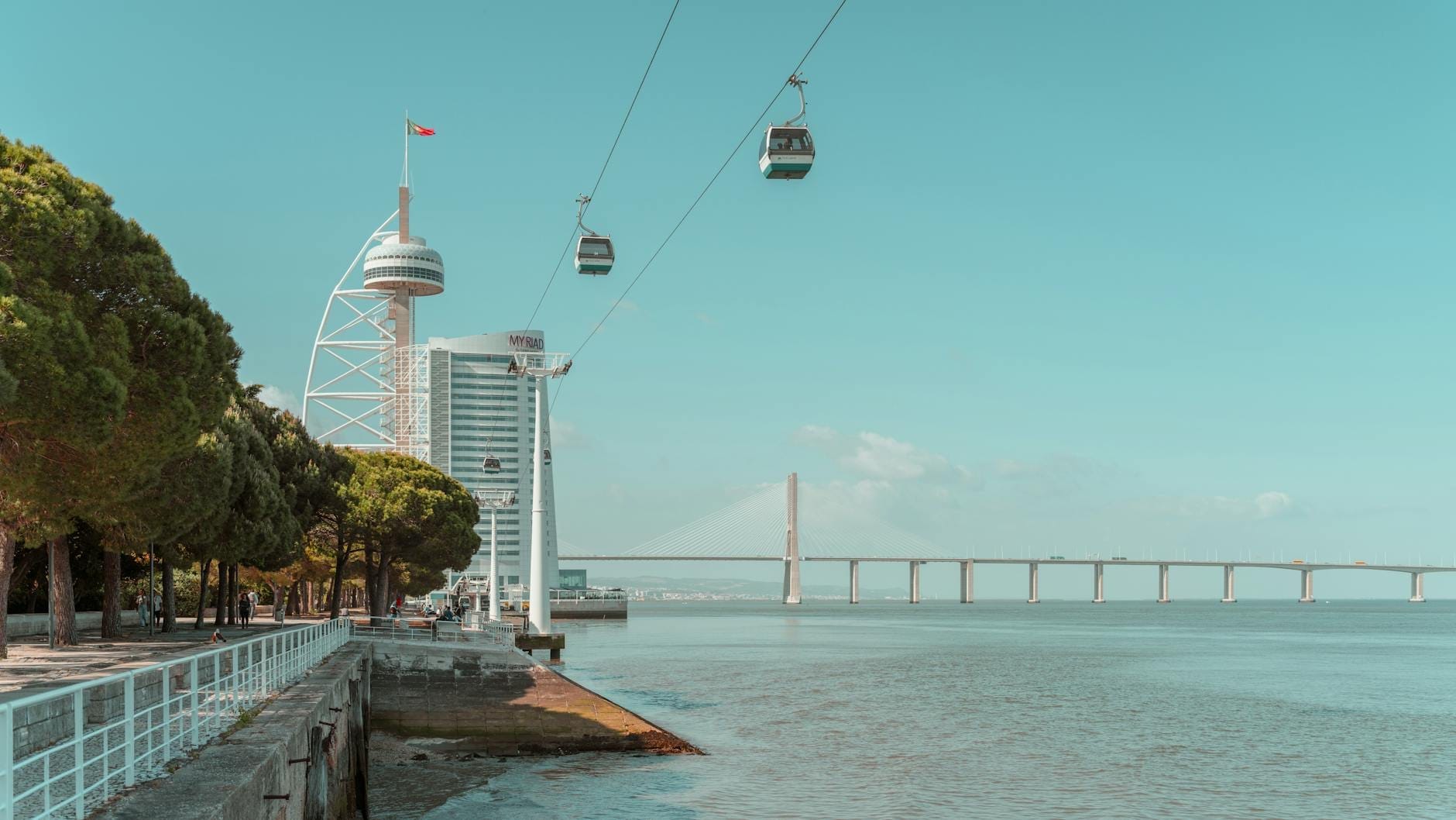 Vue sur le fleuve dans le Parque das Nações avec des bâtiments modernes, des télécabines, la tour et le pont Vasco da Gama, et une promenade bordée d'arbres.