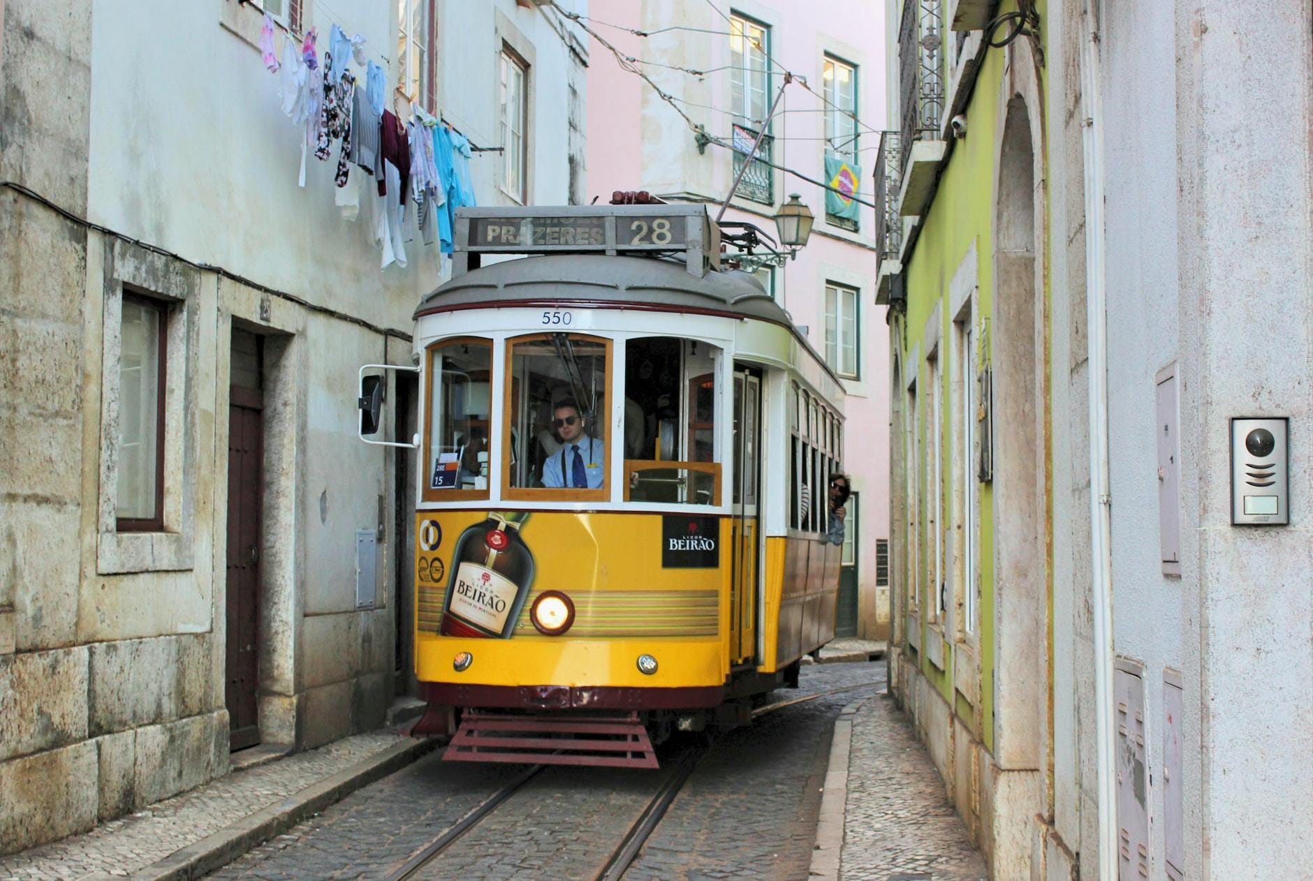 Tramway jaune vintage numéro 28 circulant dans une rue étroite du quartier historique de Lisbonne avec des bâtiments aux tons pastel et du linge suspendu au-dessus.
