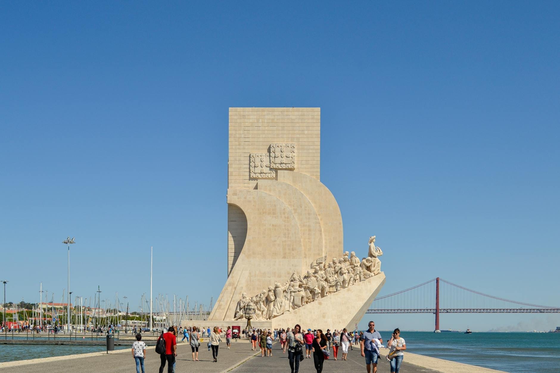 Vue large du monument Padrão dos Descobrimentos de Lisbonne le long des berges avec des promeneurs et le Pont 25 de Abril en arrière-plan.