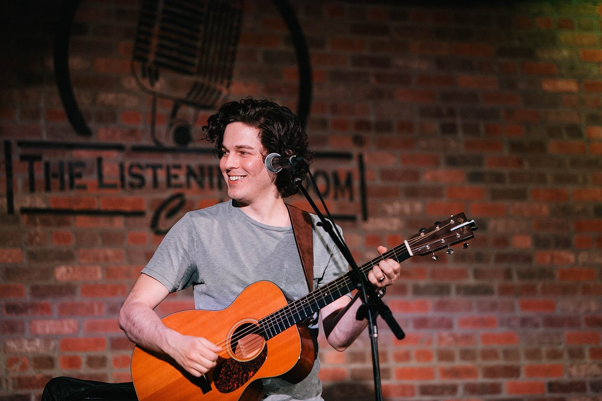 Ryan Harris Brown sonriente toca la guitarra acústica en el escenario de The Listening Room Cafe con una pared de ladrillo y el logotipo de fondo.