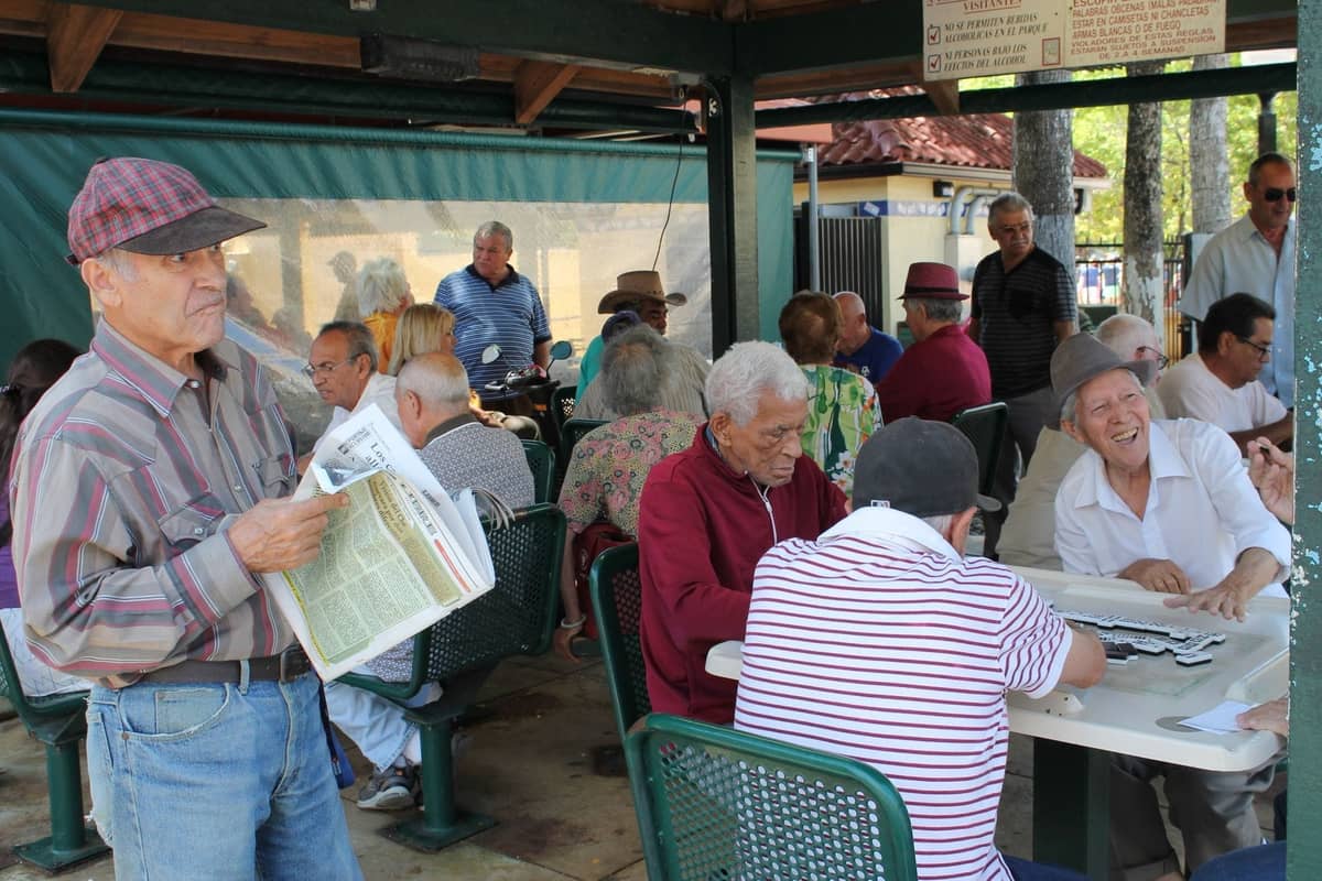 Group of elderly men playing dominoes and socializing under a shaded area in an outdoor park setting.