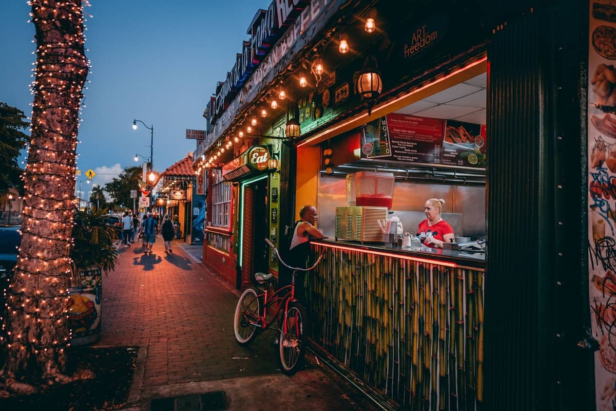 Vibrant evening view along Calle Ocho in Little Havana, Miami, with people, string lights, colorful storefronts, and a walk-up food counter.