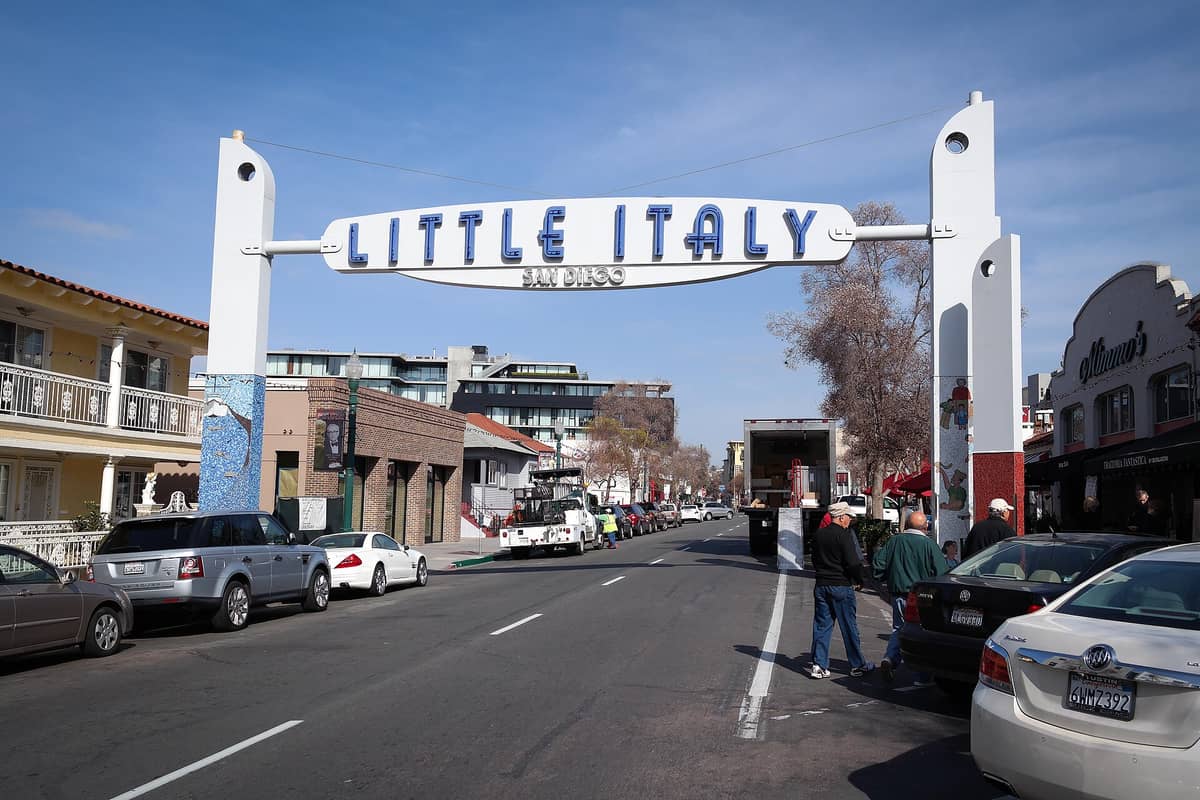 Das markante Eingangsschild von Little Italy San Diego spannt sich über die India Street – darunter Fußgänger, Autos und lokale Gebäude unter strahlend blauem Himmel.