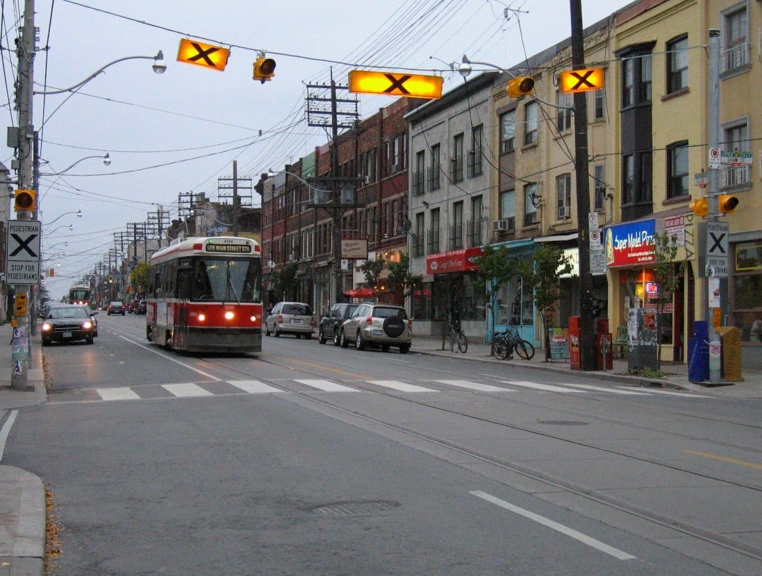 รถรางแล่นไปตาม College Street ใน Little Italy เมือง Toronto ท่ามกลางร้านค้าสีสันสดใส นักเดินถนน ท้องฟ้าครึ้ม และสัญญาณไฟจราจร