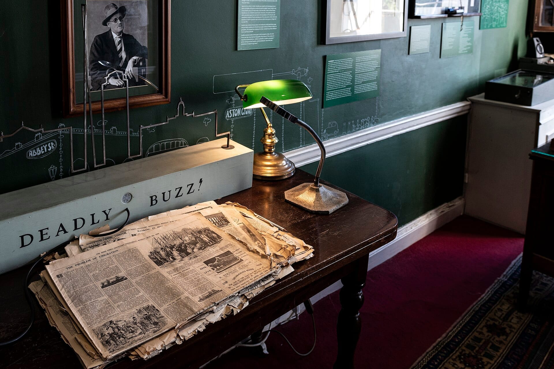 Un'esposizione al Little Museum of Dublin con vecchi giornali, una lampada da banchiere verde e manufatti storici su una scrivania in legno scuro.