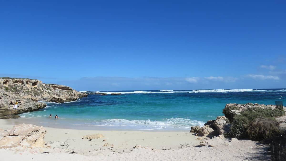 Little Salmon Bay auf Rottnest Island mit türkisfarbenem Wasser, Sandstrand, felsigen Klippen und einigen schwimmenden Menschen unter einem klaren blauen Himmel.