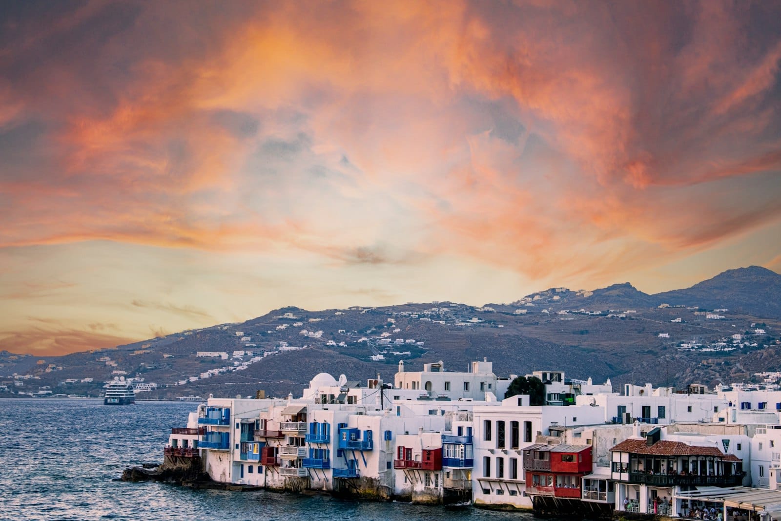 Casas costeras encaladas con coloridos balcones en Little Venice, Mykonos, bajo un cielo de atardecer dramático sobre el mar Egeo y las colinas al fondo.