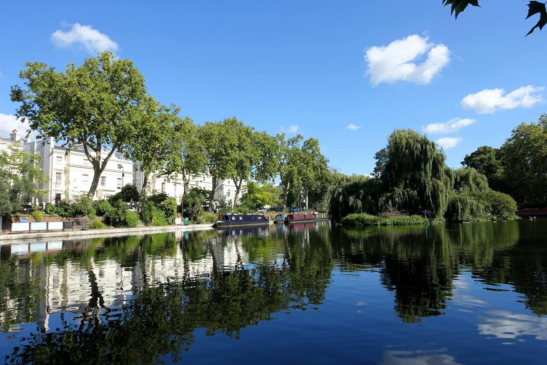 Vista panoramica delle acque tranquille di Little Venice, Londra, con chiatte ormeggiate, alberi rigogliosi e eleganti case bianche in stile Regency riflesse nell'acqua in una giornata di sole.
