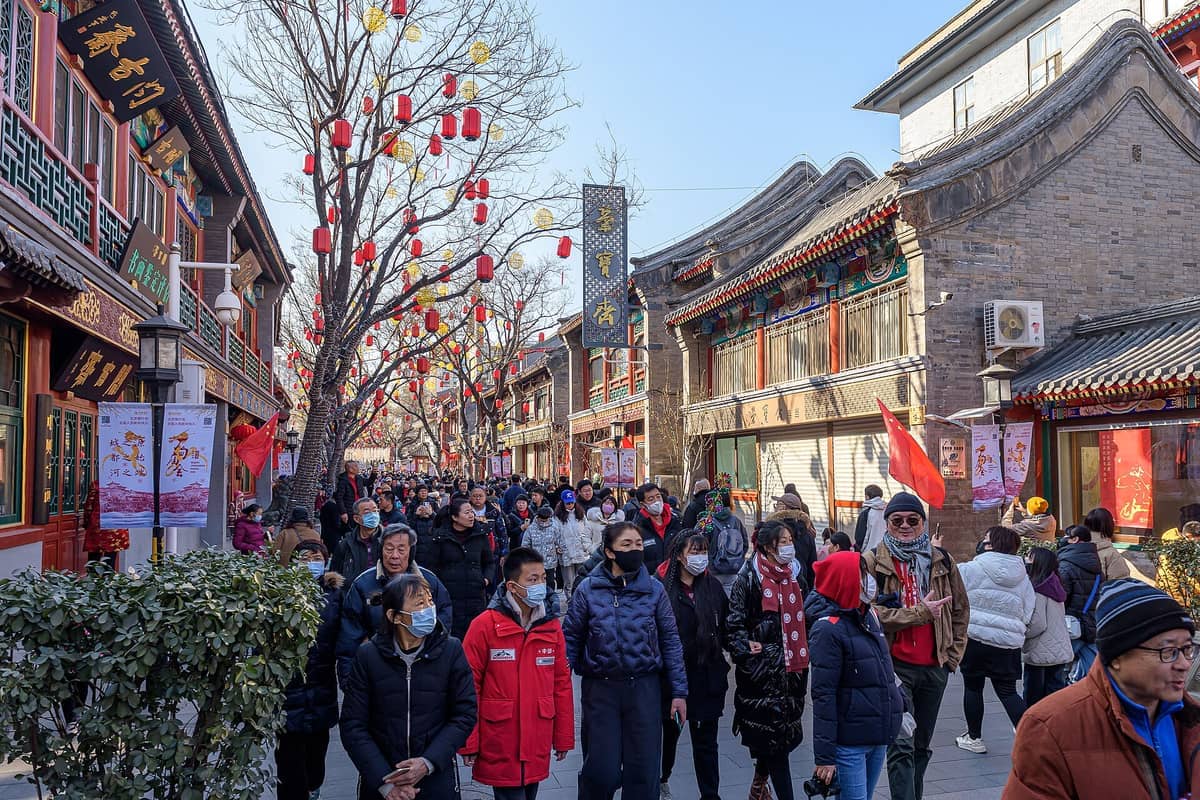 Menschenmassen schlendern entlang der Kulturstraße Liulichang, gerahmt von traditionellen Ladenfront der Qing-Dynastie und roten Laternen an einem sonnigen Tag.