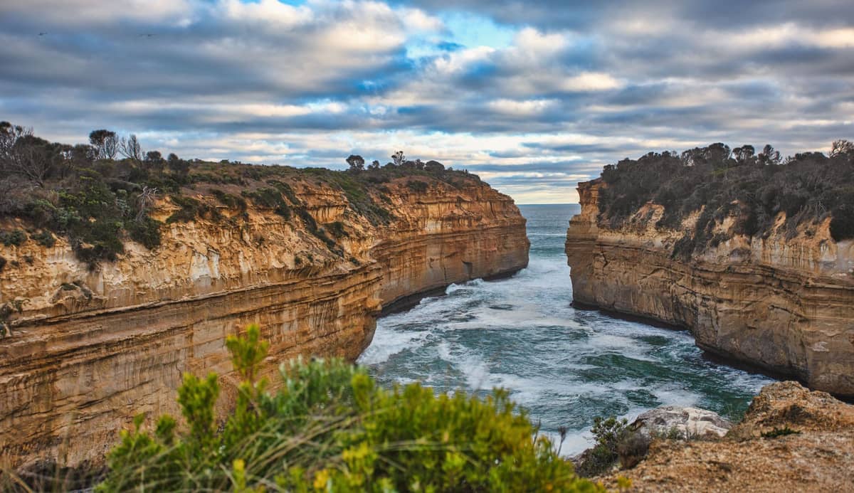 Vista aérea de los dramáticos acantilados de arenisca de Loch Ard Gorge y el océano embravecido, con la estrecha ensenada tallada por las olas y rodeada de vegetación costera.