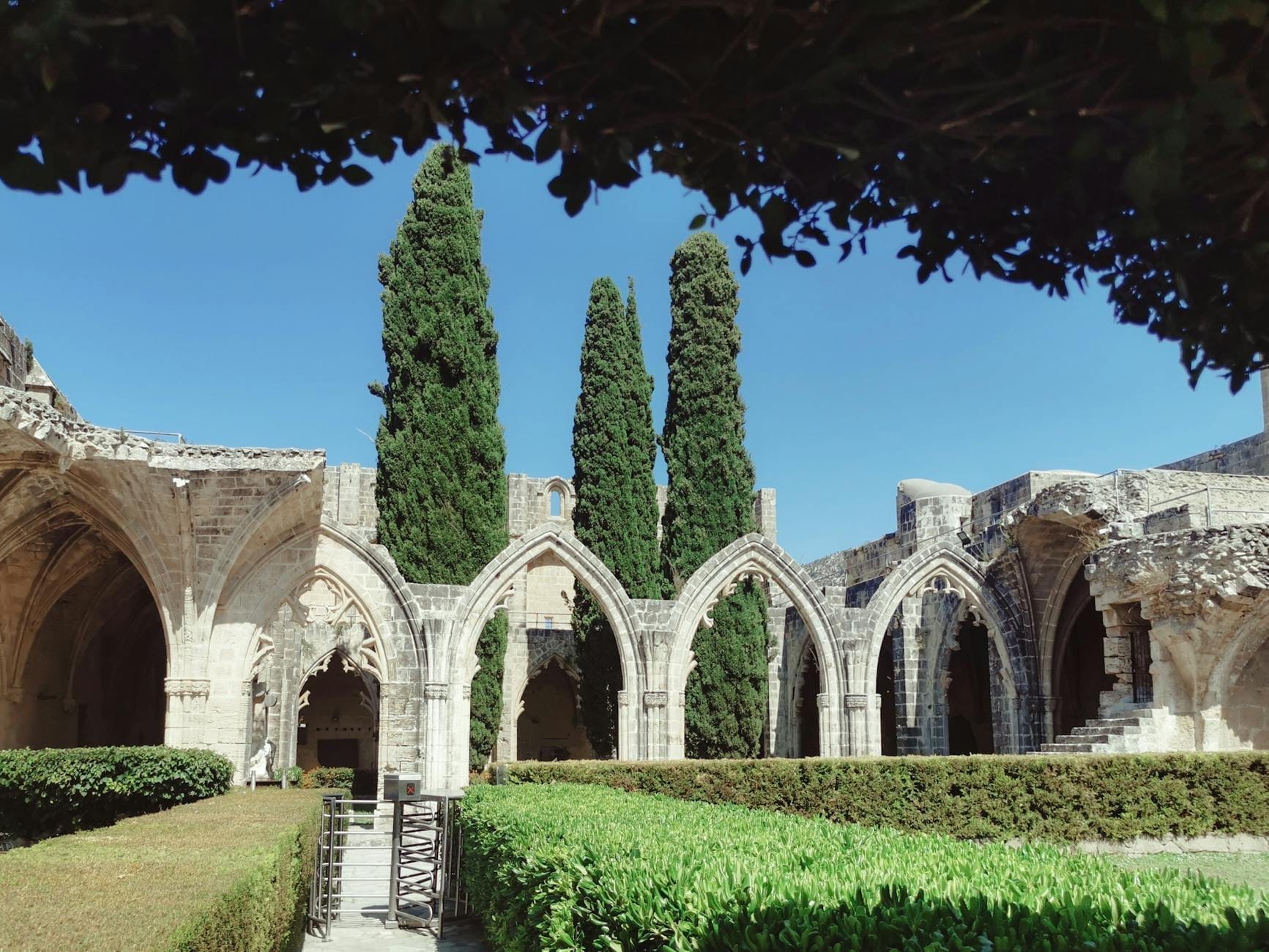 Claustros de piedra en ruinas de un monasterio medieval con arcos apuntados, rodeados de setos bien cuidados y altos cipreses bajo un cielo azul despejado.