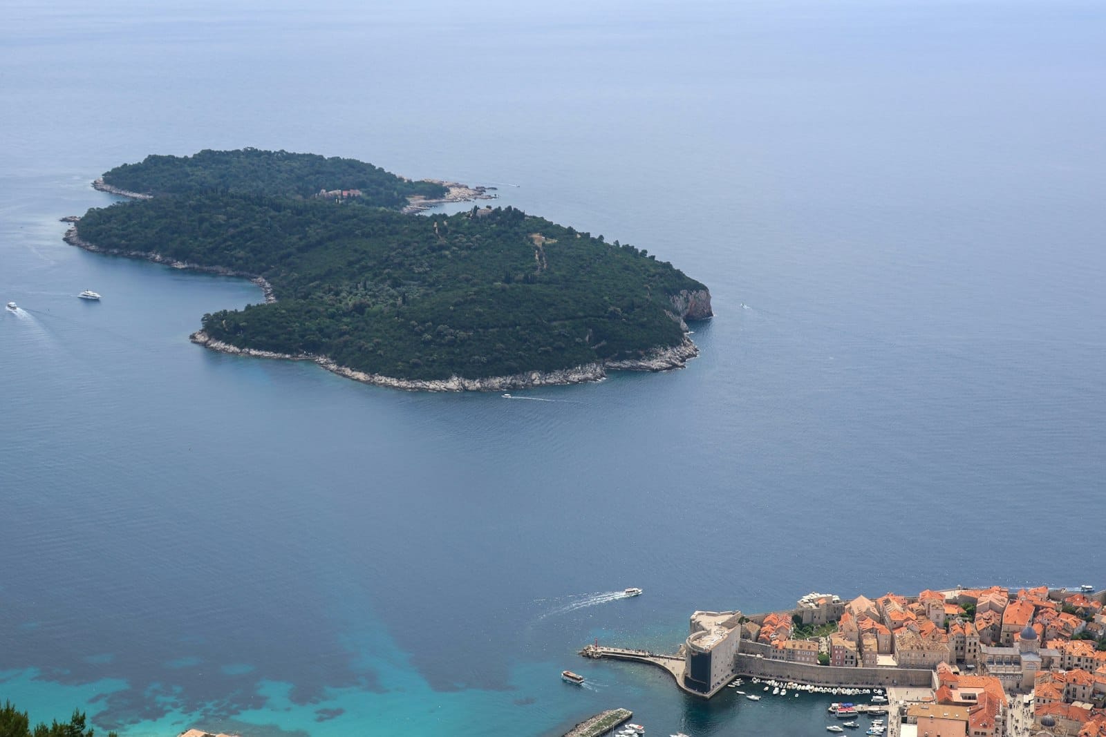 Vista aérea de la isla de Lokrum rodeada de mar azul, con la Ciudad Vieja y el puerto de Dubrovnik en primer plano en un día soleado.