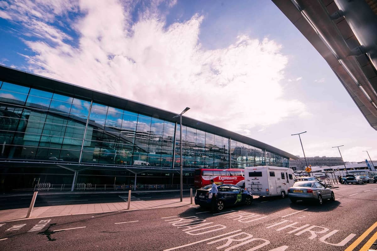 Vista exterior de um terminal de vidro moderno em um aeroporto movimentado com veículos, táxis e passageiros na frente.