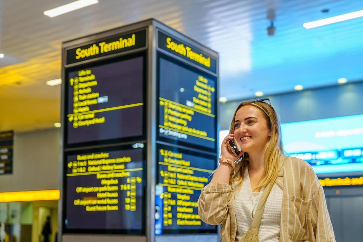Viajante sorridente ao telefone no Terminal Sul do Aeroporto de Gatwick, com painéis de informação de voos visíveis ao fundo.