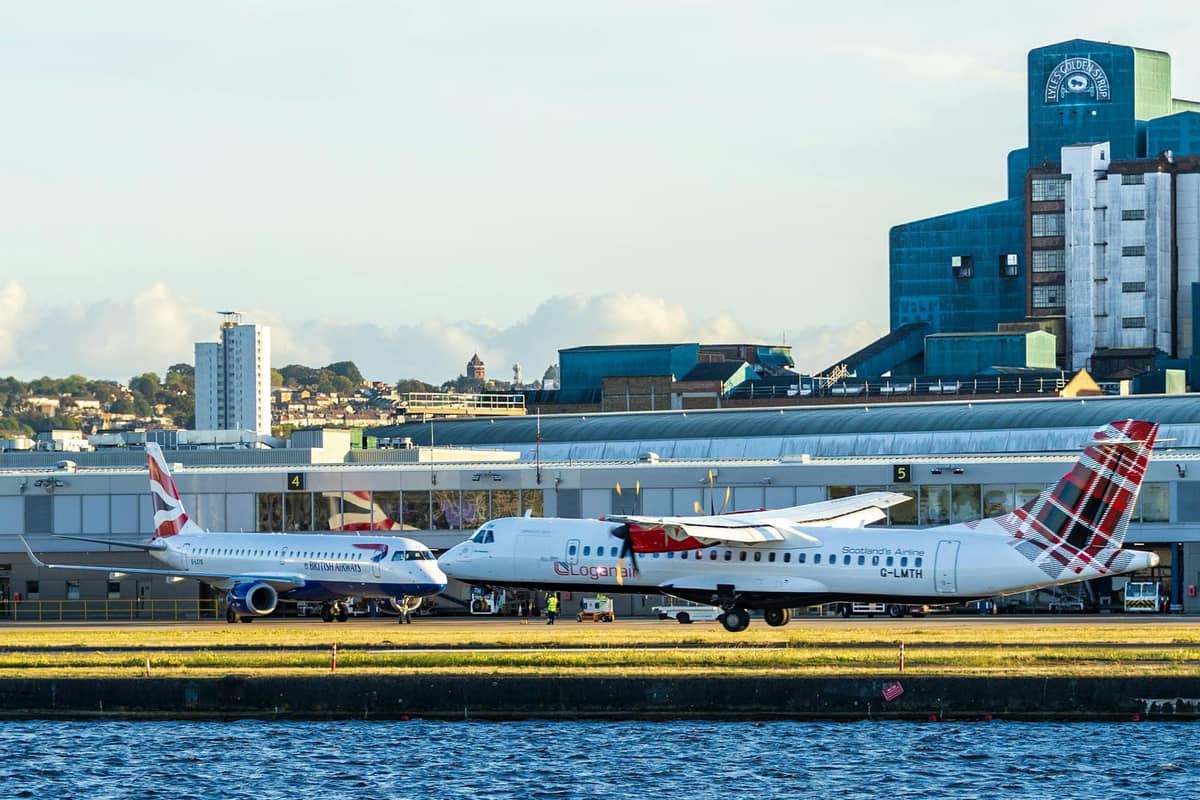 Dois aviões comerciais estacionados no London City Airport, com edifícios modernos e o terminal ao fundo, à beira da água.