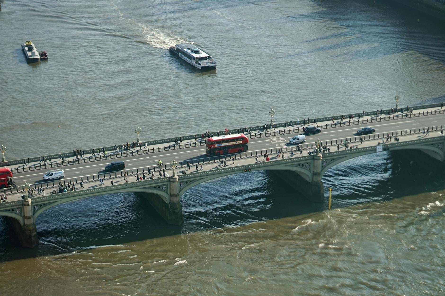 Vista aerea di London Bridge che attraversa il Tamigi con un autobus rosso a due piani, auto e pedoni, barche sul fiume e palazzi sullo sfondo.