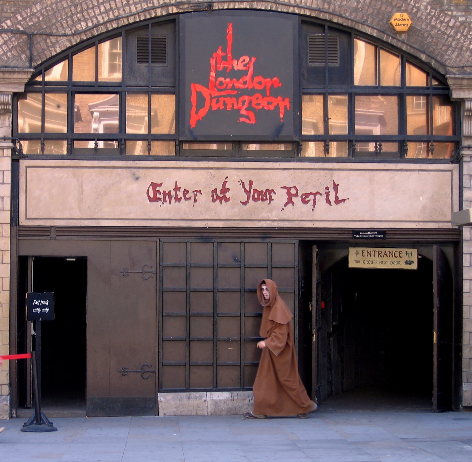 Ingresso del The London Dungeon con insegne gotiche, finestre ad arco e un attore in costume con un mantello marrone che cammina all'esterno.